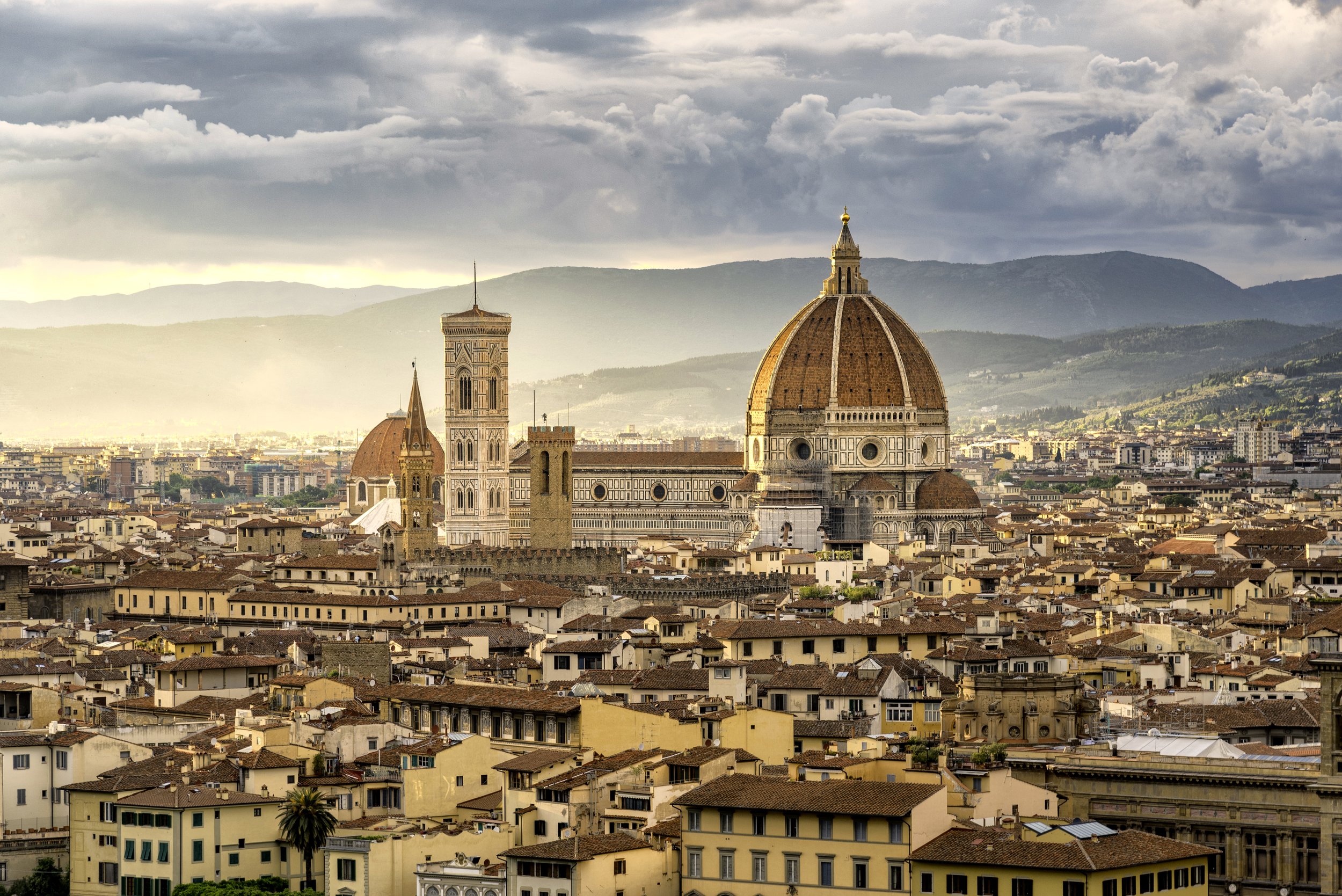 Panorama di Firenze con il Duomo di Firenze, la Cupola di Brunelleschi e le torri medievali, con montagne sullo sfondo e nuvole sopra.