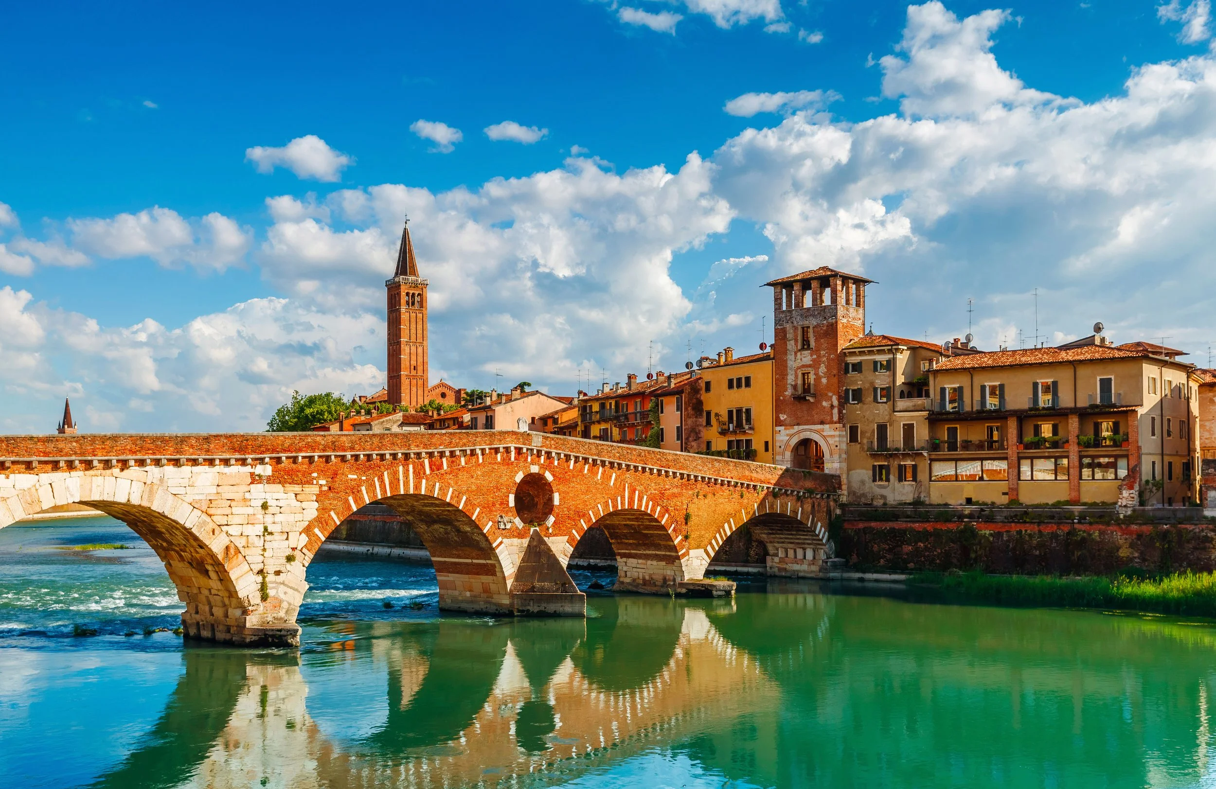 Un ponte in pietra con archi sotto un cielo nuvoloso, con edifici storici e una torre campanaria in lontananza.