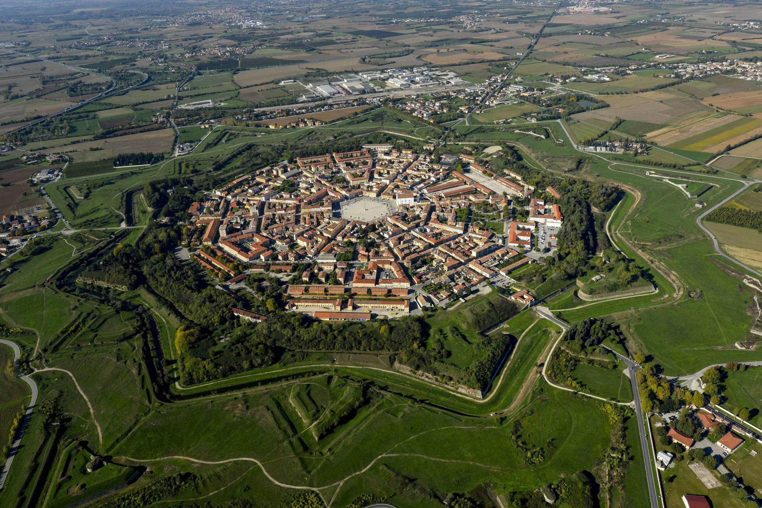 Veduta aerea di un centro storico circondato da campi verdi e strade, con edifici in cotto e alcune mura medievali visibili.