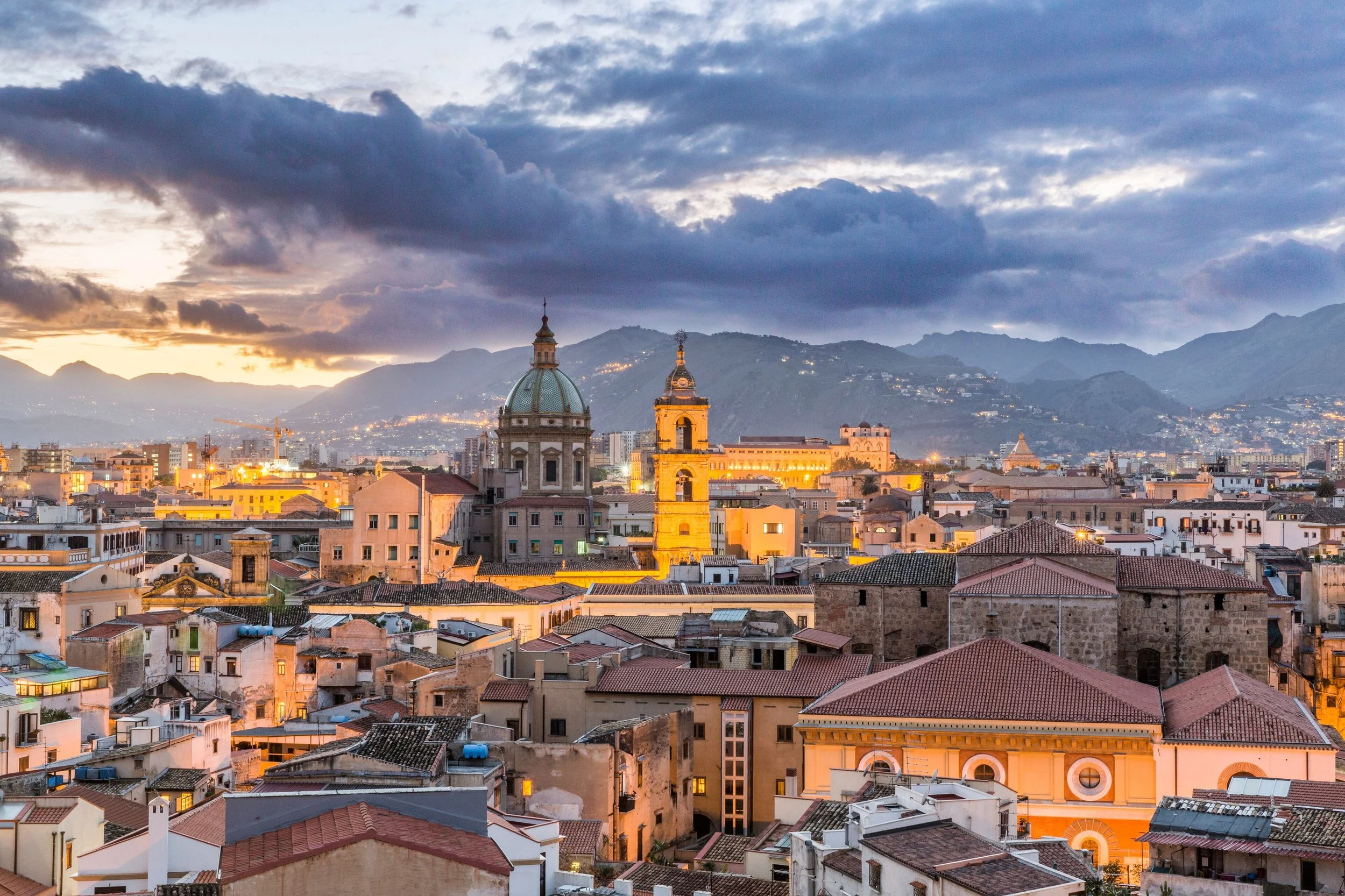 Volo panoramico di una città con chiese storiche e montagne sullo sfondo, al tramonto, con il cielo nuvoloso e luci che illuminano gli edifici.