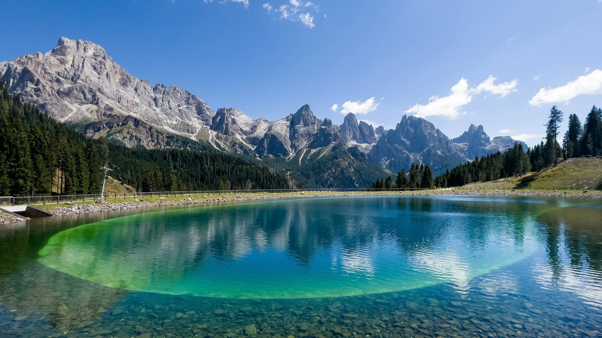 Lago di montagna circondato da alberi verdi e catene montuose rocciose con cielo blu e nuvole sparse.