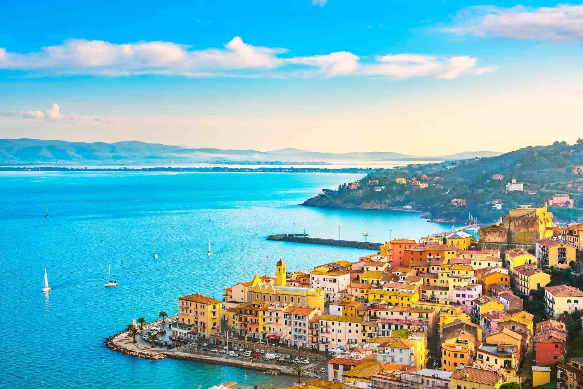Vista panoramica di un piccolo porto con case colorate lungo la costa, sullo sfondo il mare e le colline.
