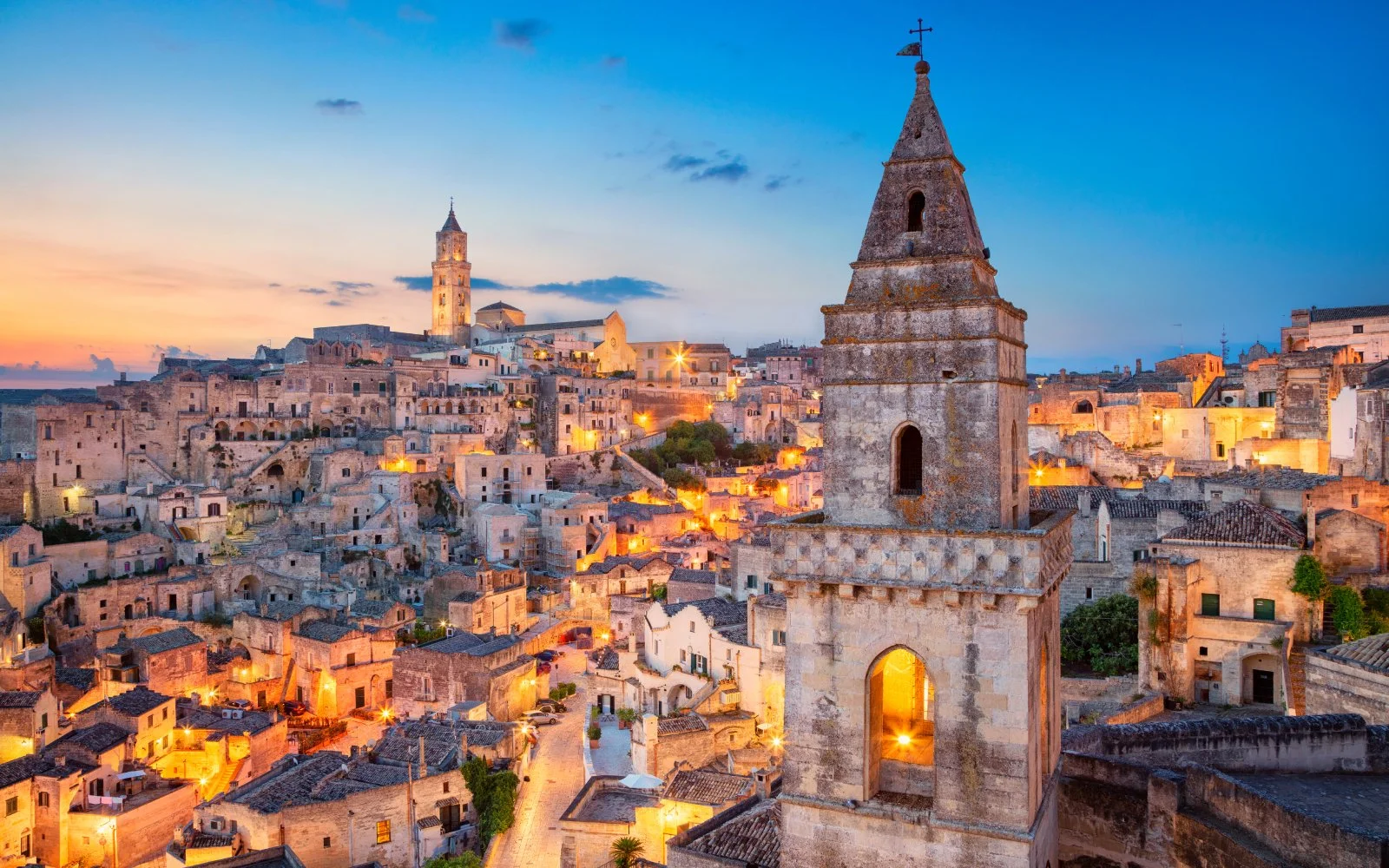 Paesaggio urbano di una città antica con edifici in pietra, campanile in primo piano e chiese storiche sullo sfondo durante il tramonto.