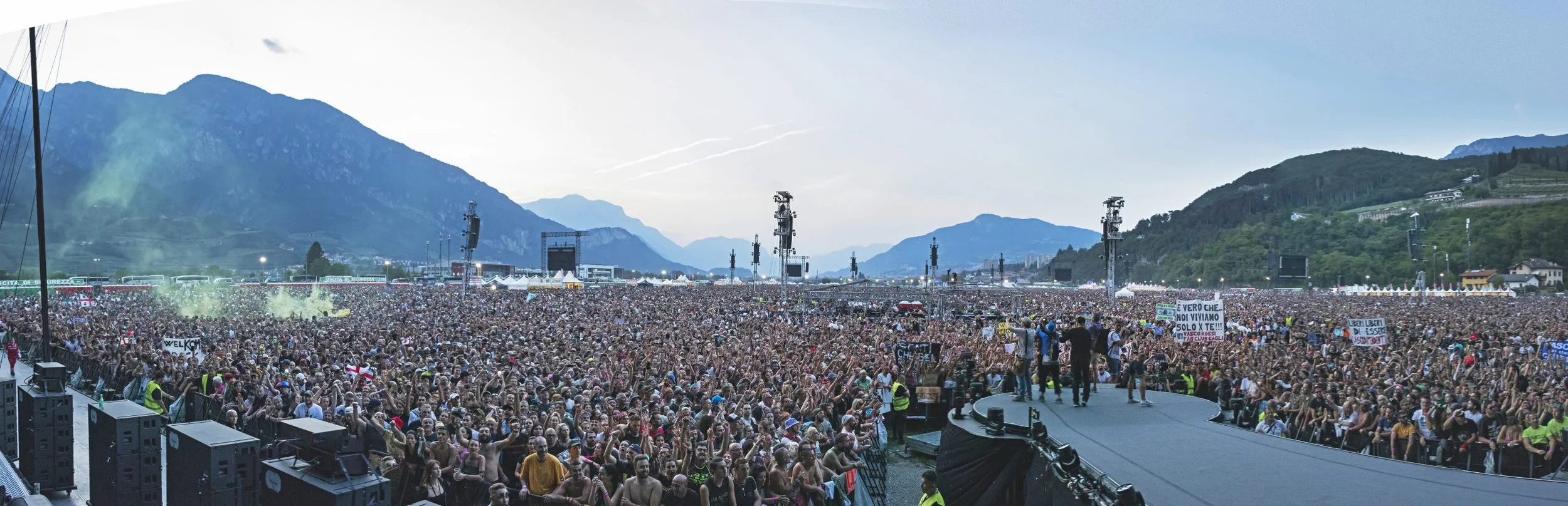Grande concerto all'aperto con migliaia di persone di fronte a un palco con montagna sullo sfondo.