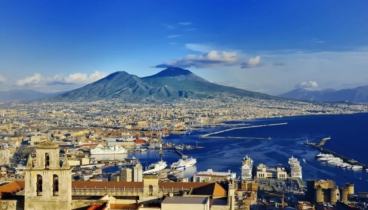 Veduta di un porto con navi da crociera, con una città sullo sfondo e un vulcano attivo, il Monte Vesuvio, che emette una piccola quantità di vapori.