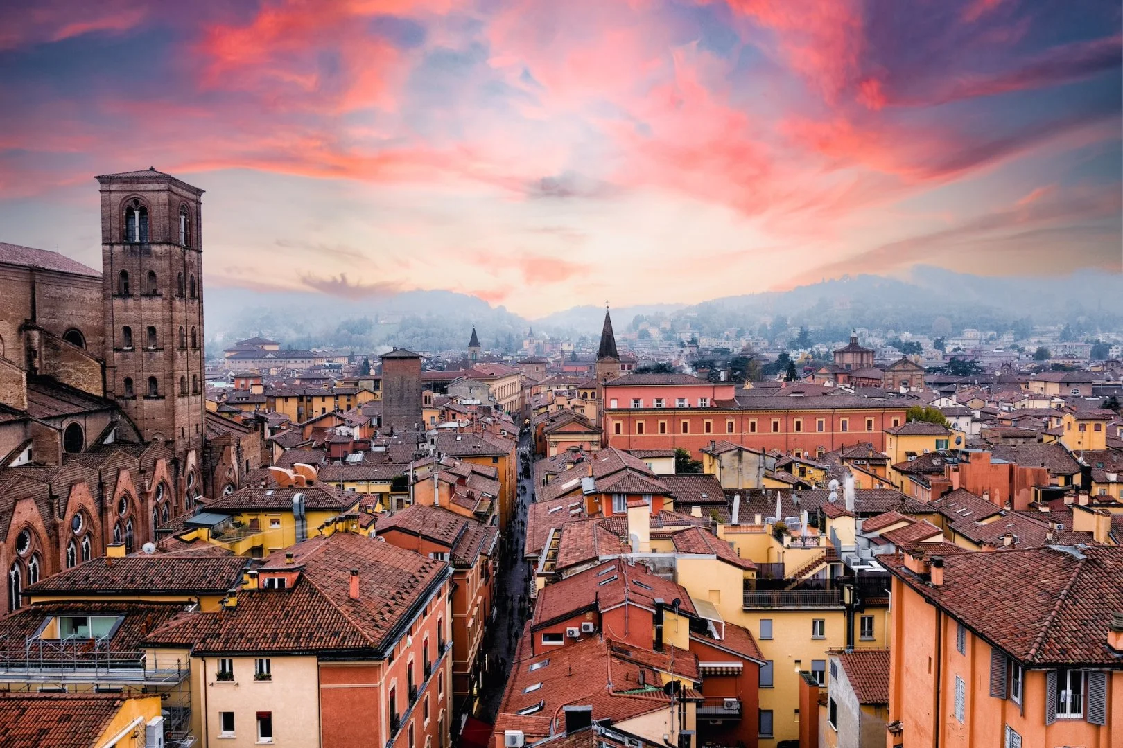 Scorcio di una città con edifici colorati e tetti di tegole, chiese con torri e un cielo al tramonto con nuvole rosa e arancioni.