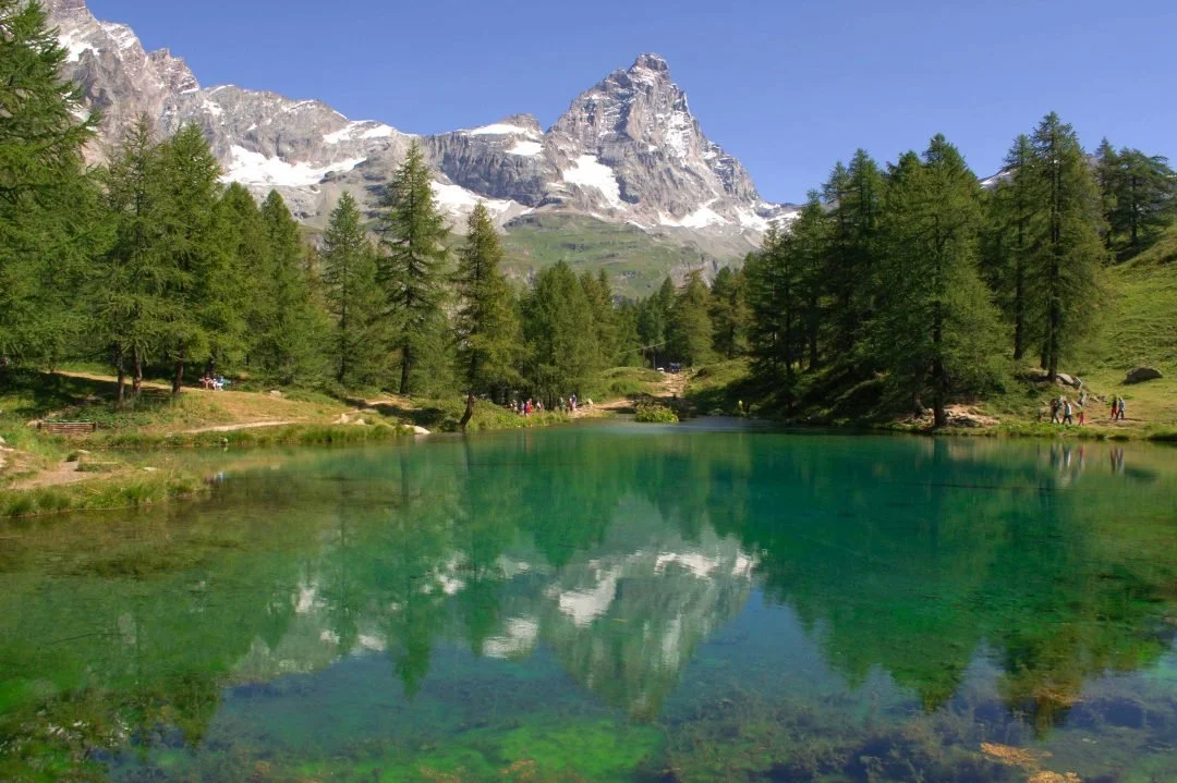 Lago di montagna con acque verdi e montagne innevate sullo sfondo, circondato da pini e sentieri con alcune persone.
