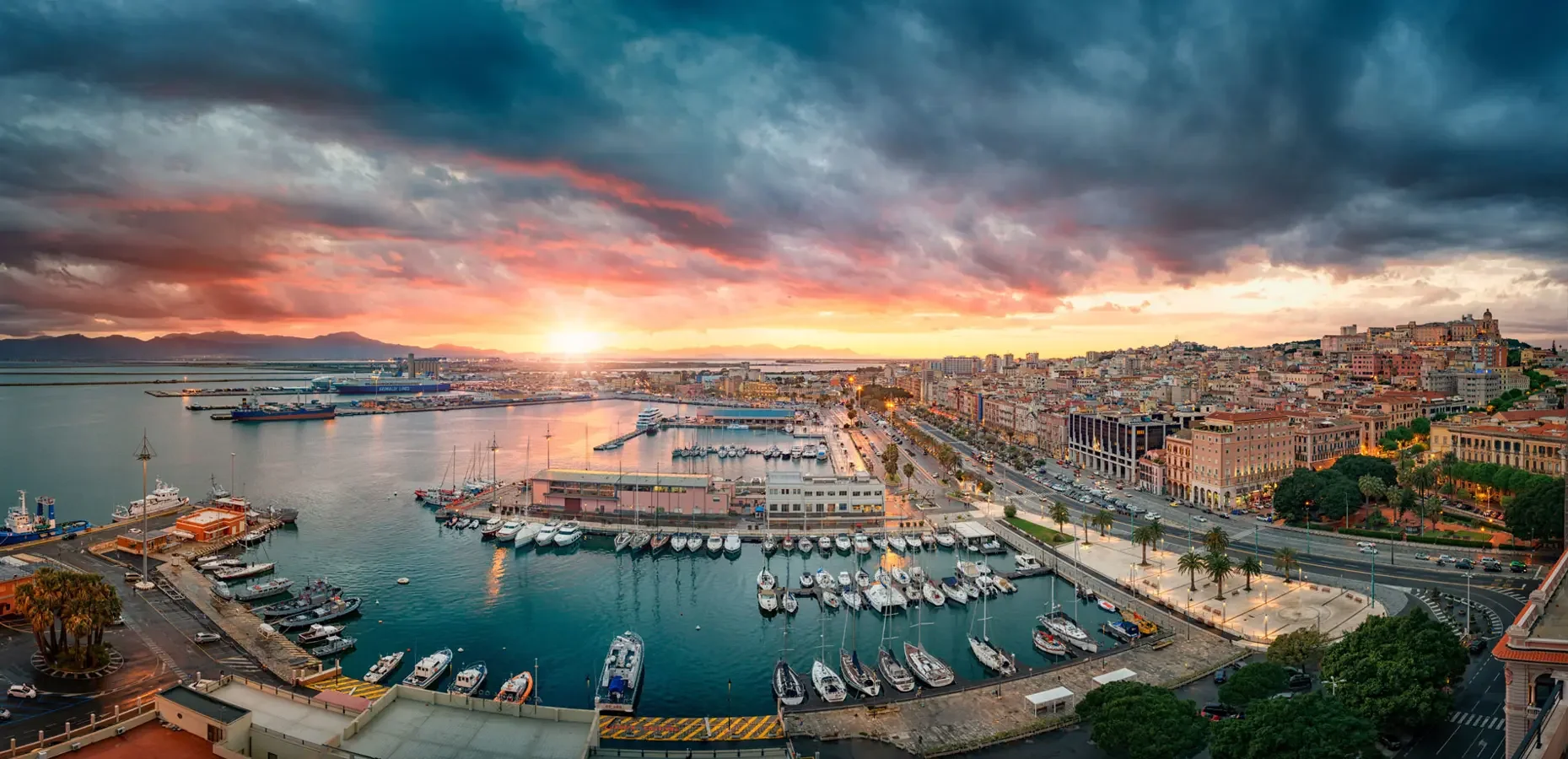 Veduta di un porto con barche e yacht, sullo sfondo una città con edifici storici e moderni, al tramonto con nuvole colorate nel cielo.