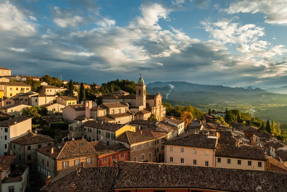Paesaggio di un borgo italiano con case e una chiesa con campanile, su colline verdi con montagne all'orizzonte e un cielo nuvoloso