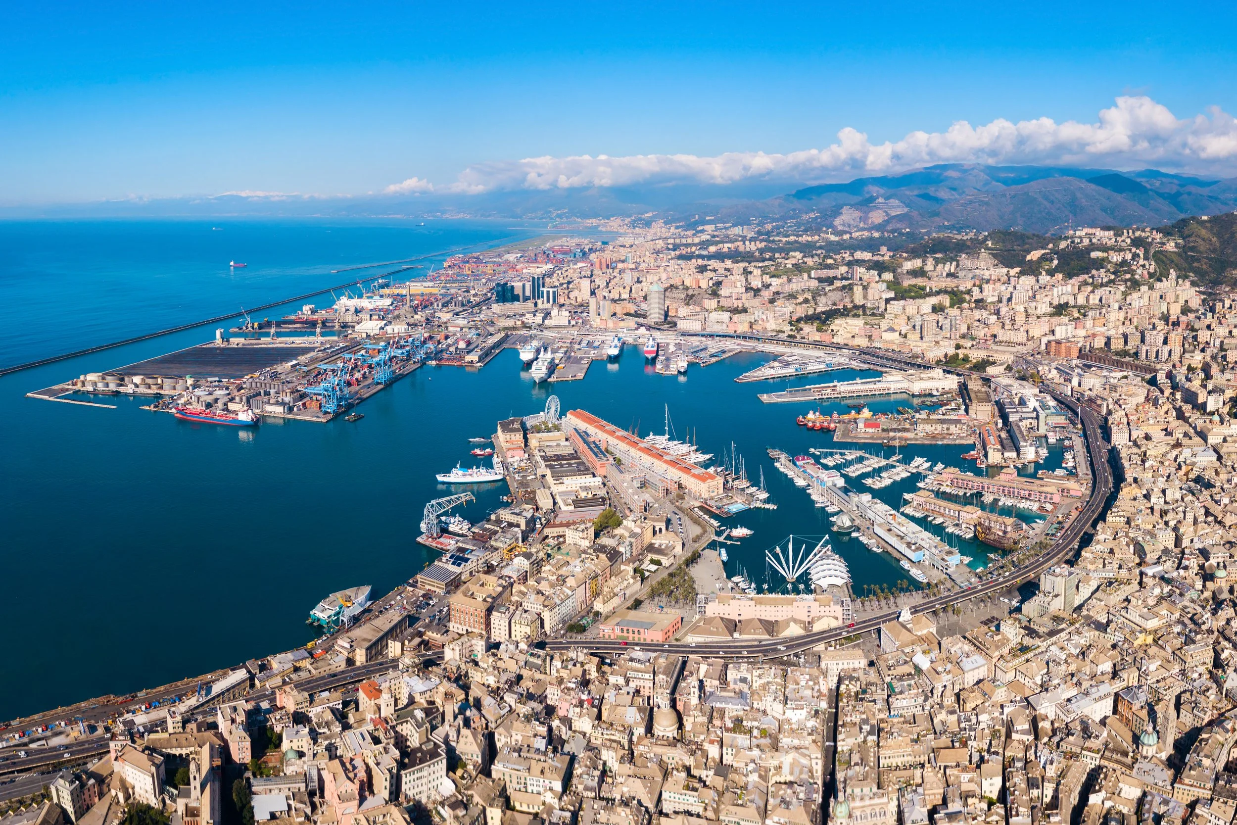 Vista aerea di un porto con navi, costruzioni e strade, con montagne sullo sfondo e cielo sereno.
