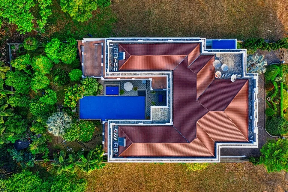 Veduta aerea di una grande casa con tetto rosso, piscina rettangolare blu e giardini verdi circostanti.