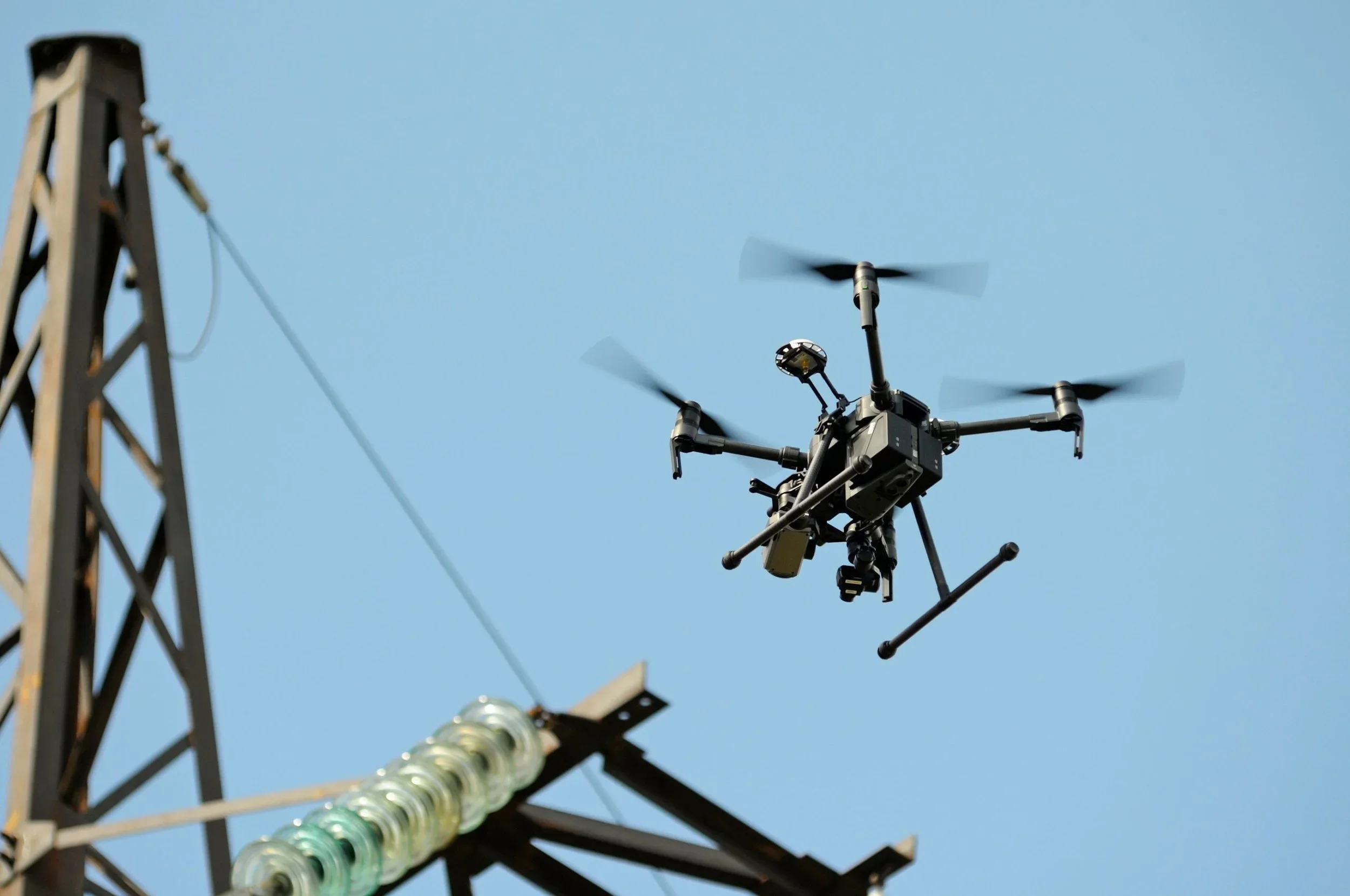 Un drone di grandi dimensioni si trova in volo vicino a una torre di supporto alle linee elettriche, sotto un cielo azzurro.