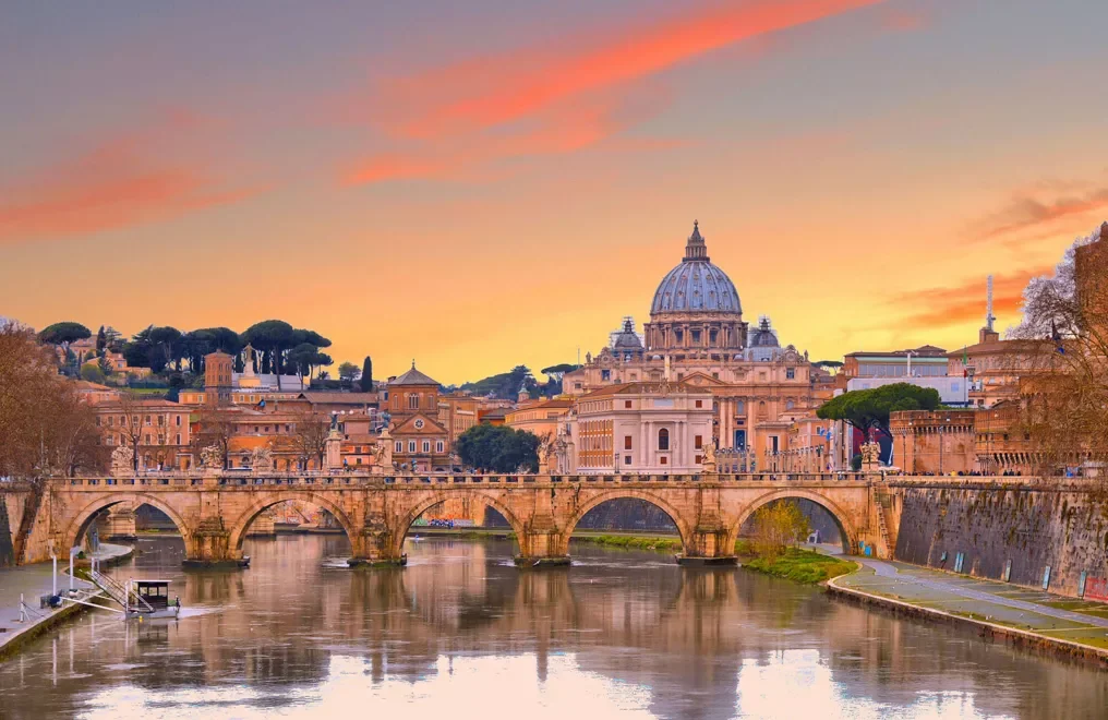 Scorcio di Roma al tramonto con il ponte Sant'Angelo sul fiume Tiber e la cupola di San Pietro sullo sfondo.