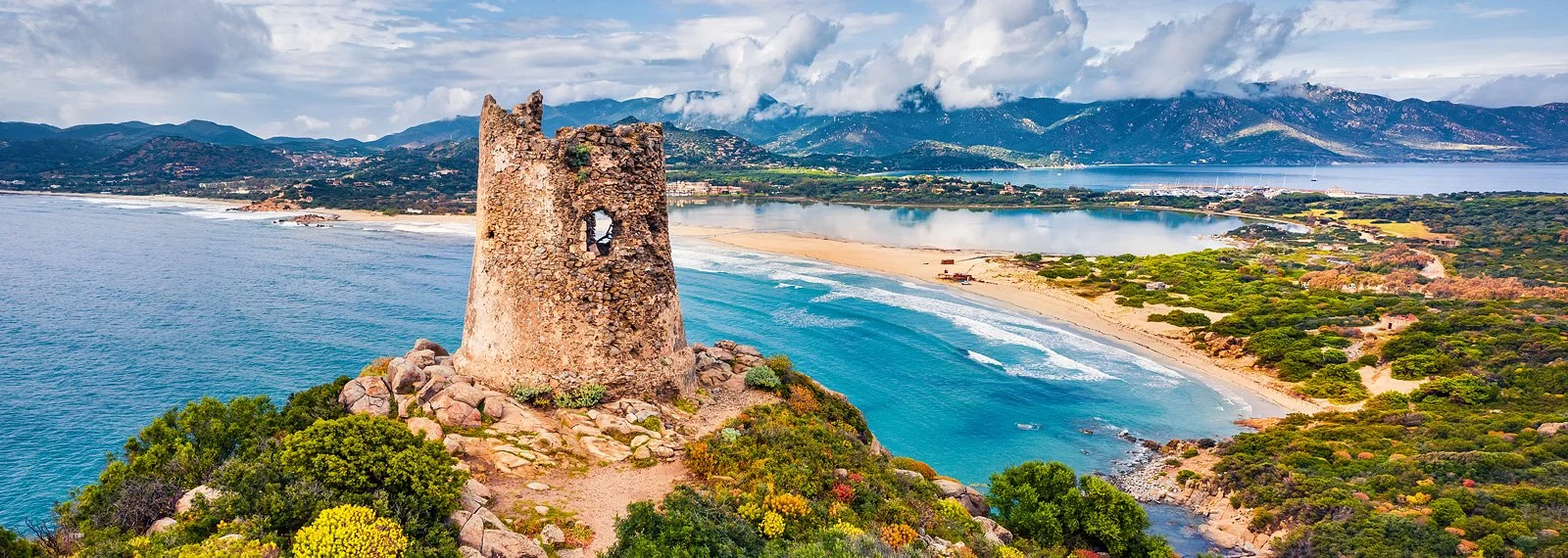 Torre diroccata sulla costa con mare, spiaggia e montagne sullo sfondo