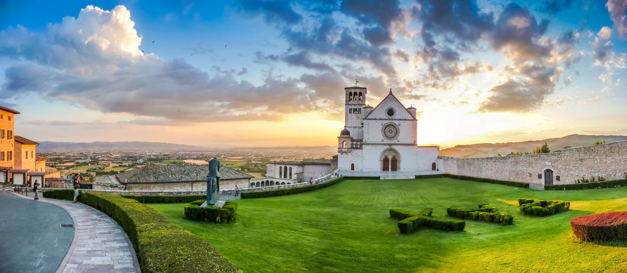 Vista del tramonto su una cattedrale bianca in cima a una collina con un ampio prato verde curato, statue e una mura di pietra, con un cielo nuvoloso e alcune persone che camminano o scattano foto.