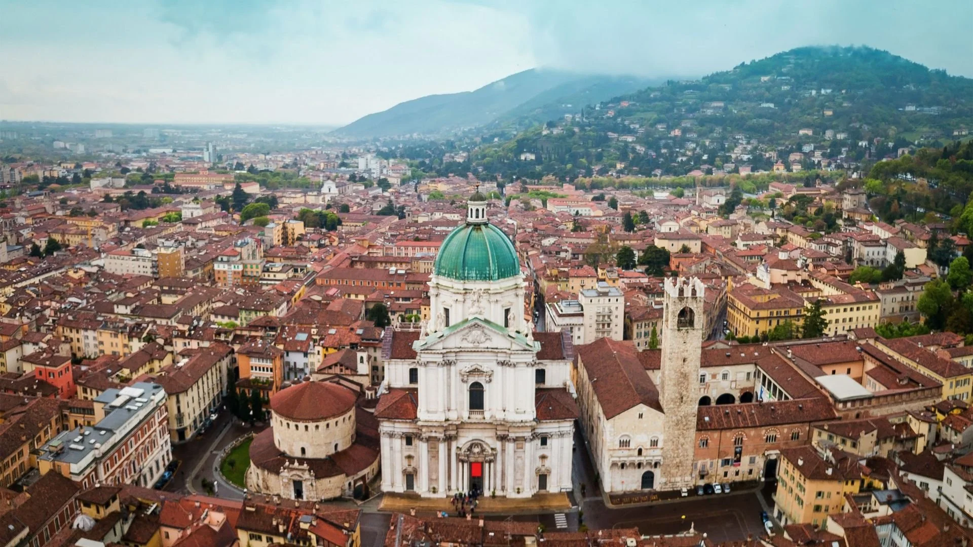 Vista panoramica di una città con una grande cattedrale con cupola verde e torre antica, circondata da edifici storici e un paesaggio montano sullo sfondo.