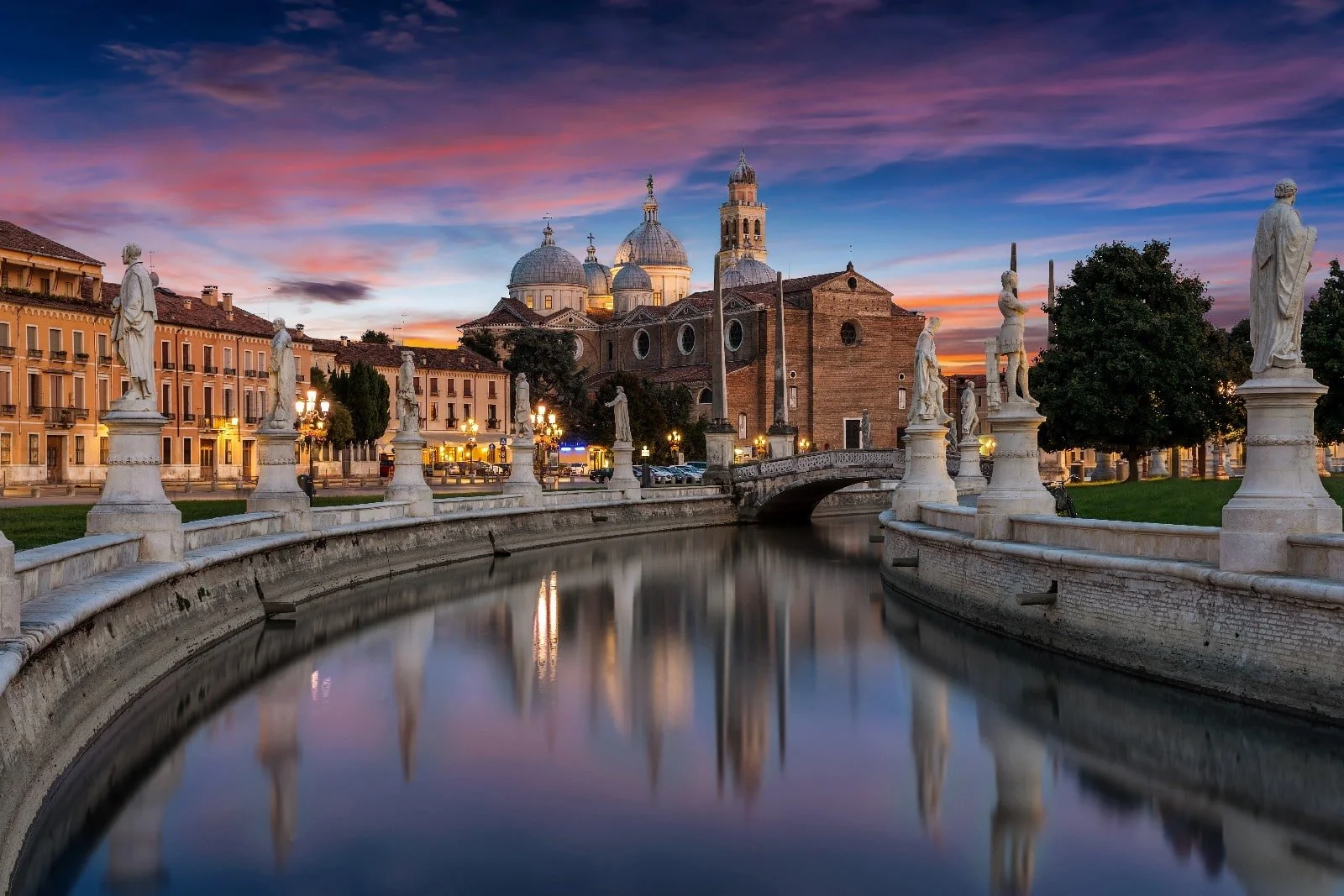 Veduta di una città con chiese storiche e statue lungo un canale durante il tramonto, in Italia.