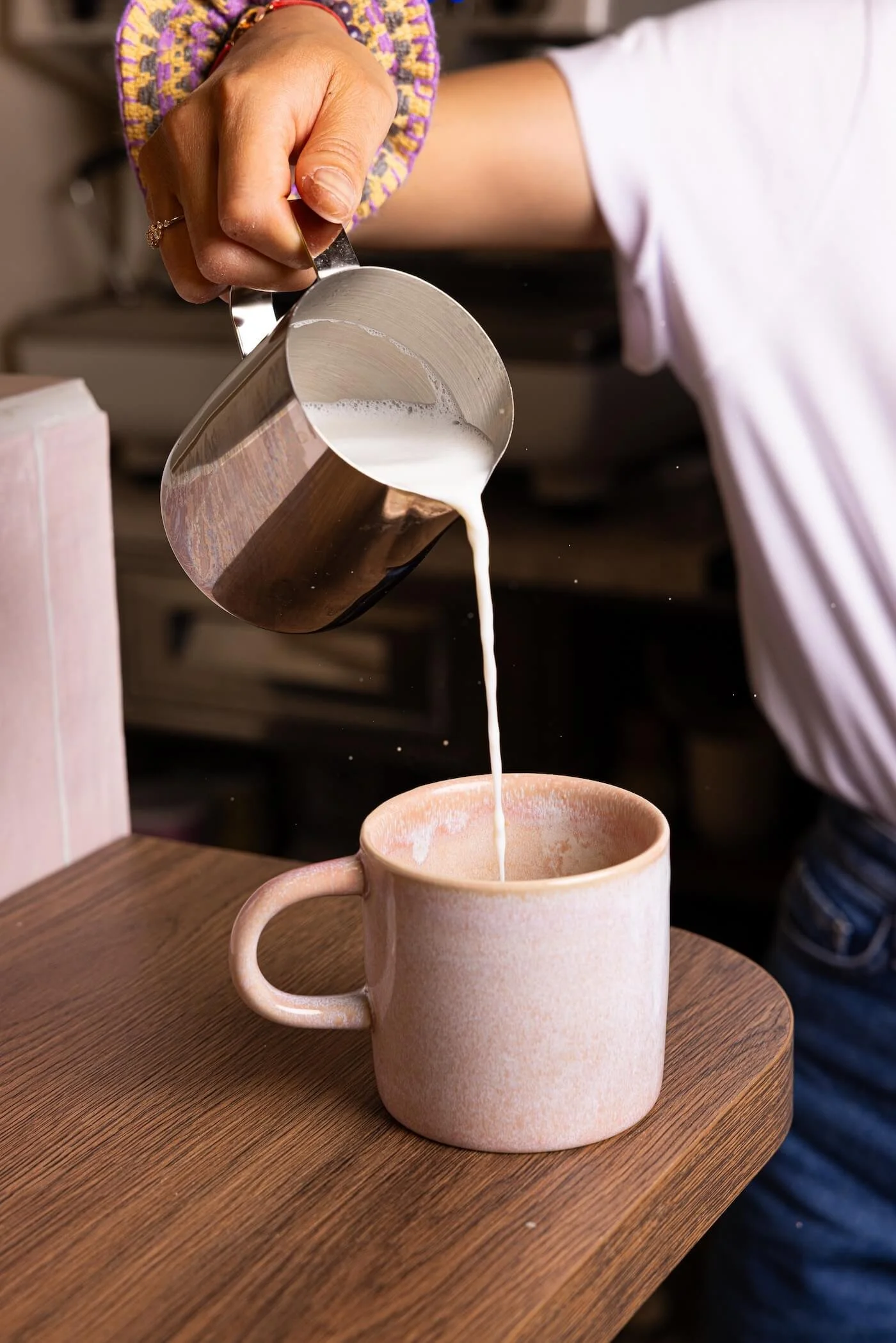 Une personne verse du lait chaud dans une tasse en céramique beige sur une assiette en bois
