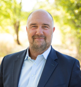 Portrait of a bald man with a beard, wearing a dark blazer and a light blue collared shirt, smiling outdoors during daytime with trees in the background.