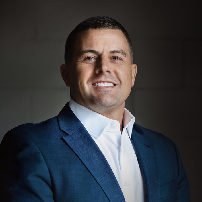 A professional headshot of a man wearing a dark blue suit and white shirt, smiling at the camera against a neutral background.