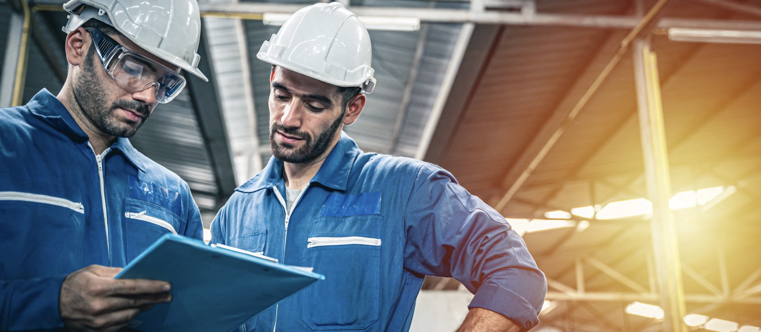 Two male workers in blue uniforms and white safety helmets looking at a clipboard inside an industrial building with metal roof structure.