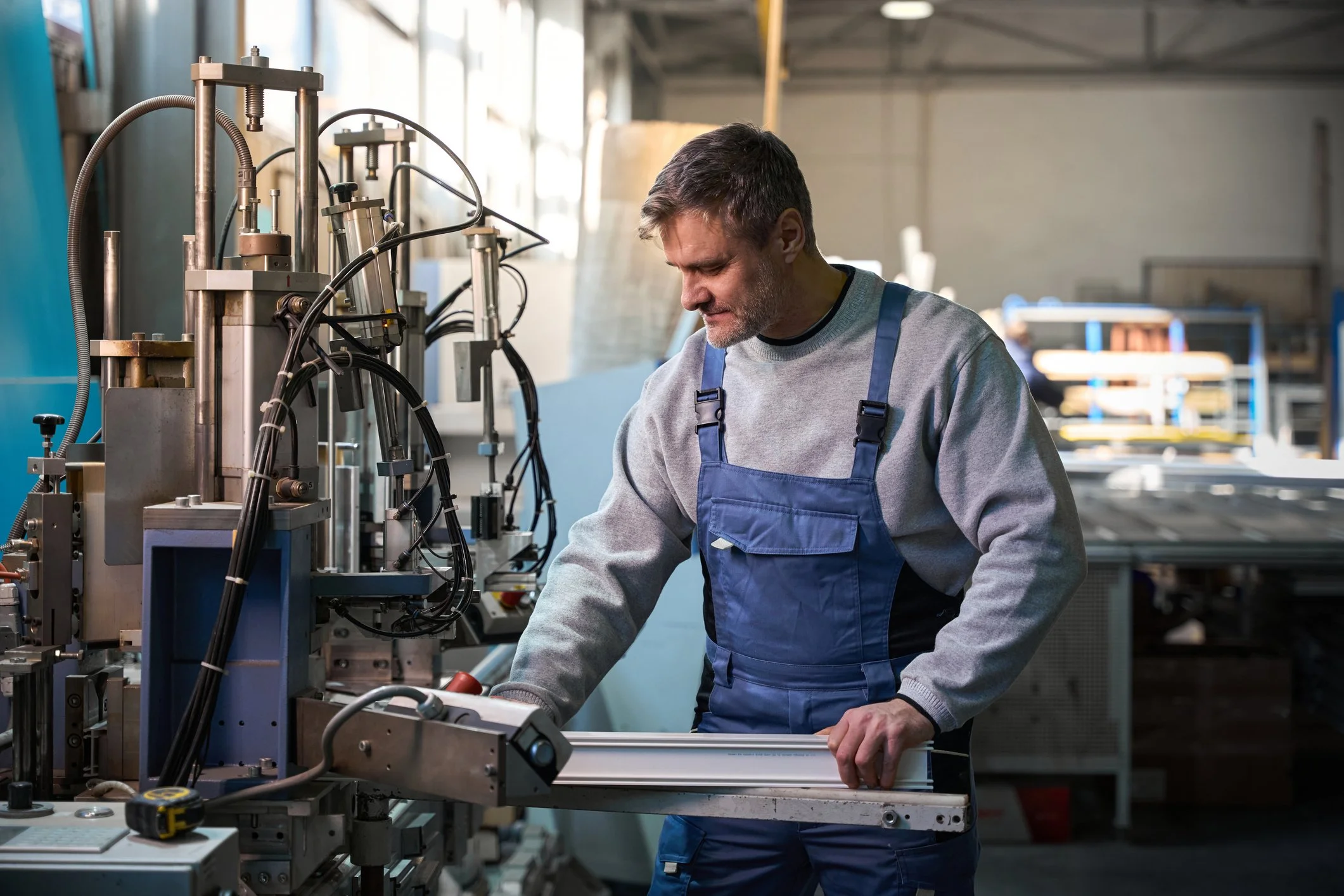 A man wearing a gray sweatshirt and blue overalls working with machinery in an industrial factory.