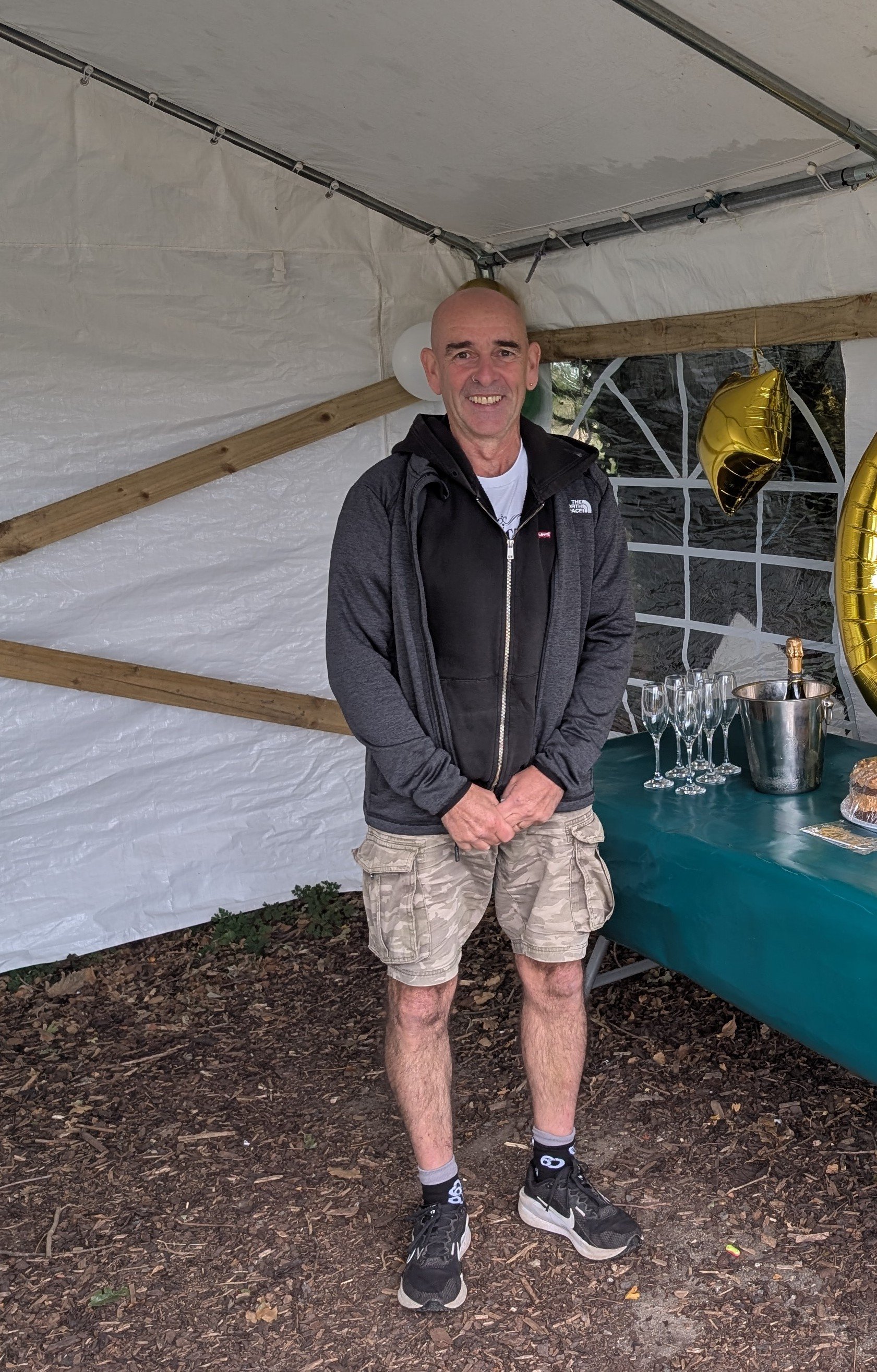 A man standing inside a decorated tent at a celebration, smiling at the camera. There are gold balloons, champagne glasses, and a cake on a table behind him.