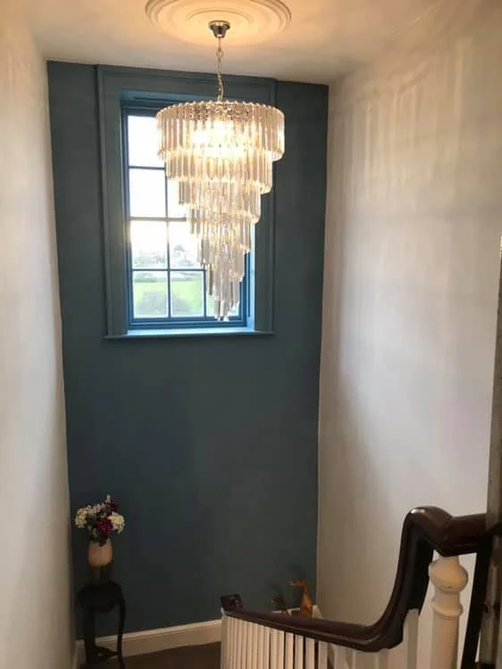 A stairwell with a blue accent wall, a window, a chandelier hanging from the ceiling, and a small floral arrangement on a black stand.