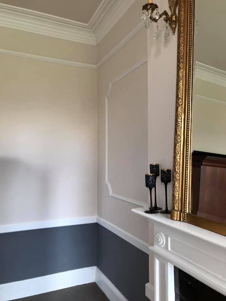 A corner of a room with beige walls and a decorative gray bottom border, white crown molding, a large gold framed mirror, a small chandelier, and black candlesticks on a white fireplace mantel.