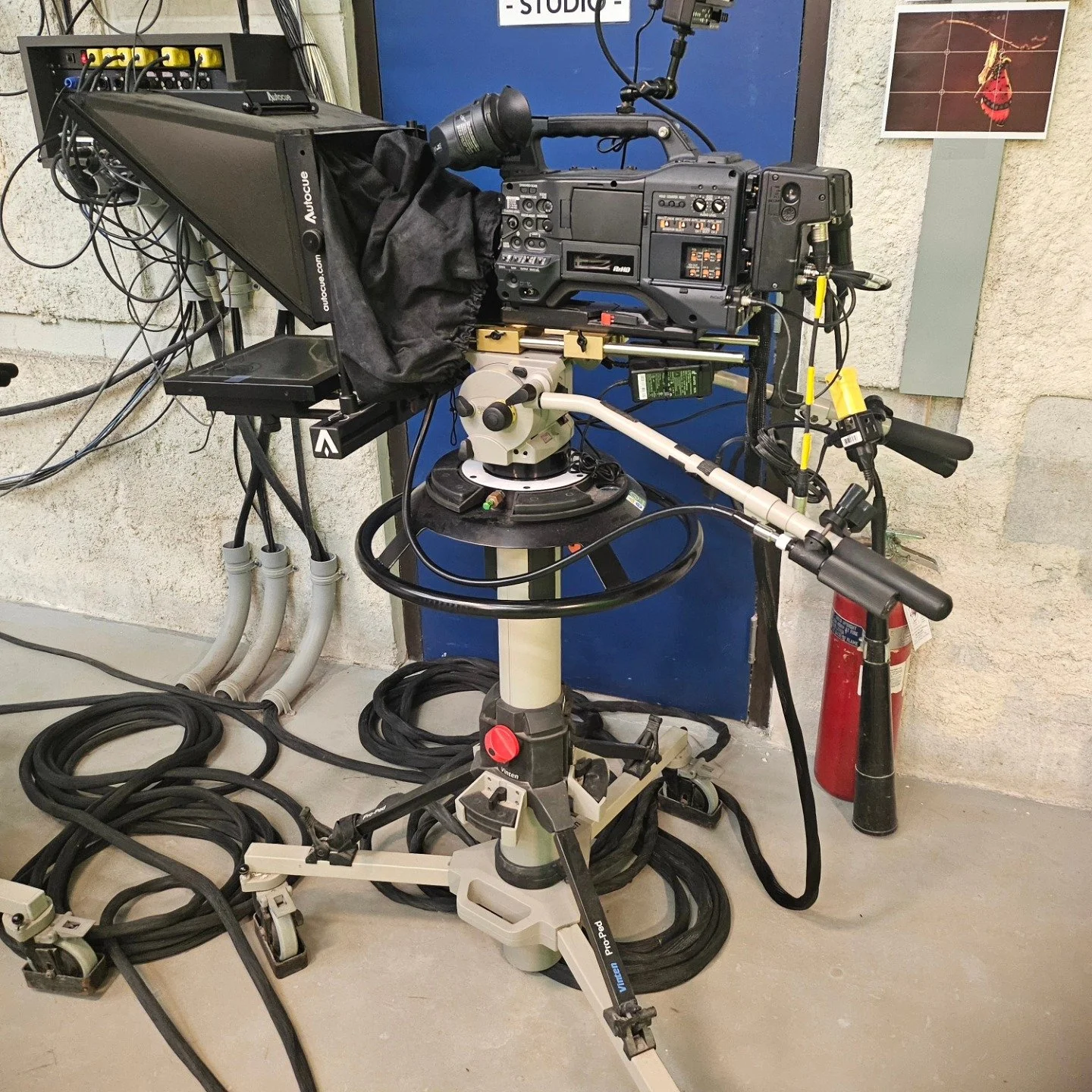 A professional video camera mounted on a tripod with various cables and equipment, set up in a studio environment.