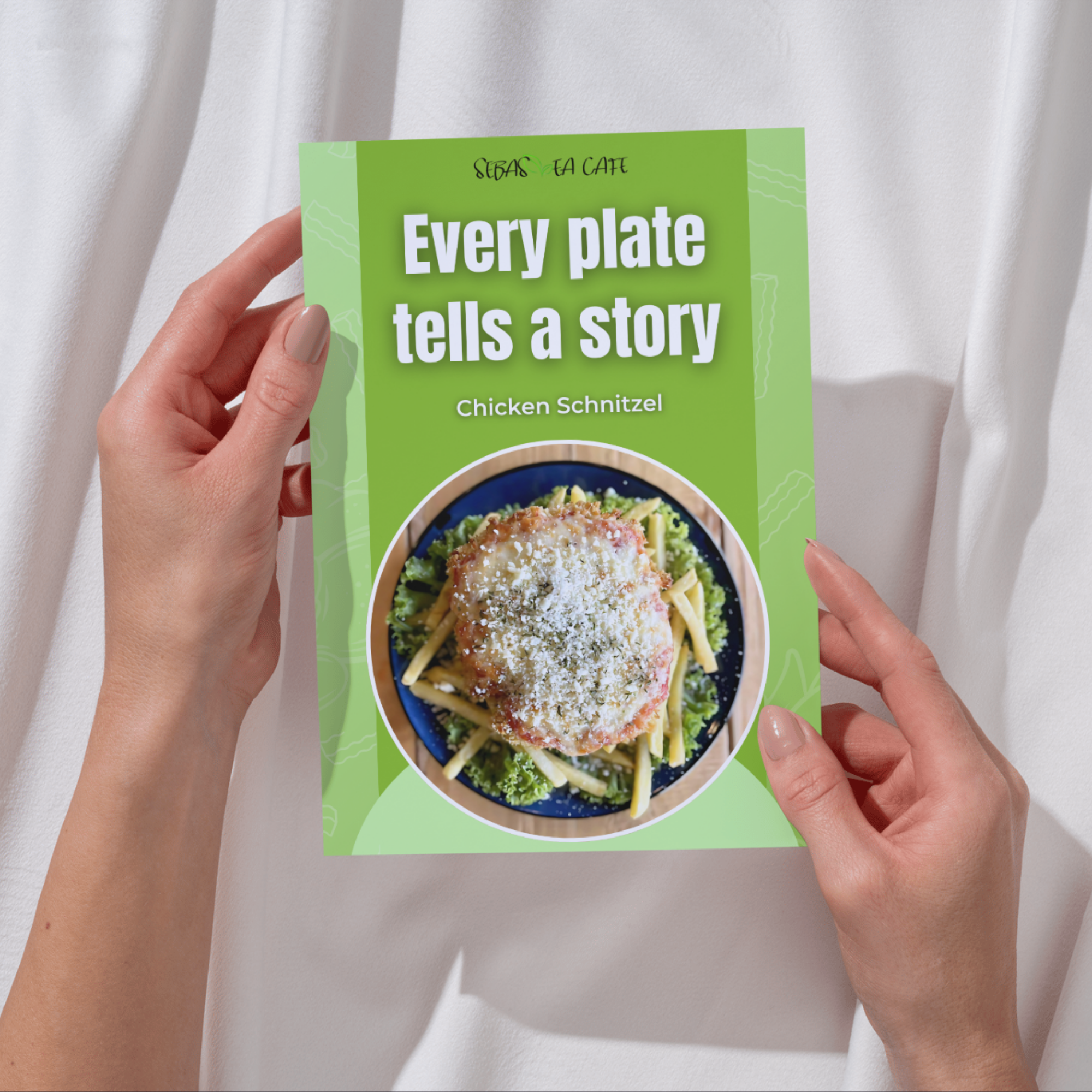A person's hands holding a green magazine or flyer with the headline 'Every plate tells a story' and a picture of a plate of chicken schnitzel with noodles and salad on a gray surface.