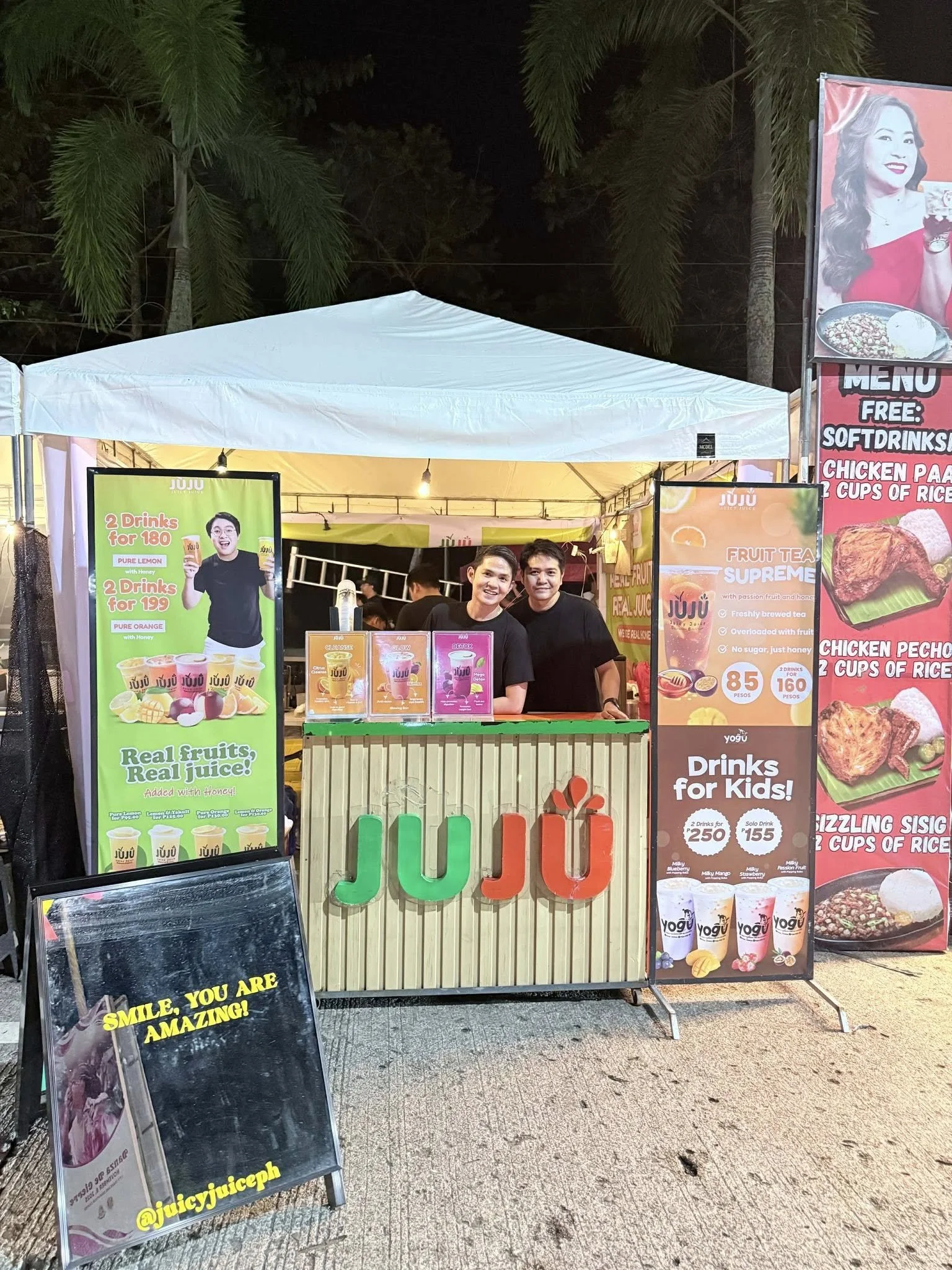 JuJu juice stand at night with posters advertising drinks, including fruit juices and smoothies, and a sign with the JuJu logo made of colorful letters. There are two people behind the counter smiling, and tall posters and signs featuring images of food and drink offers.