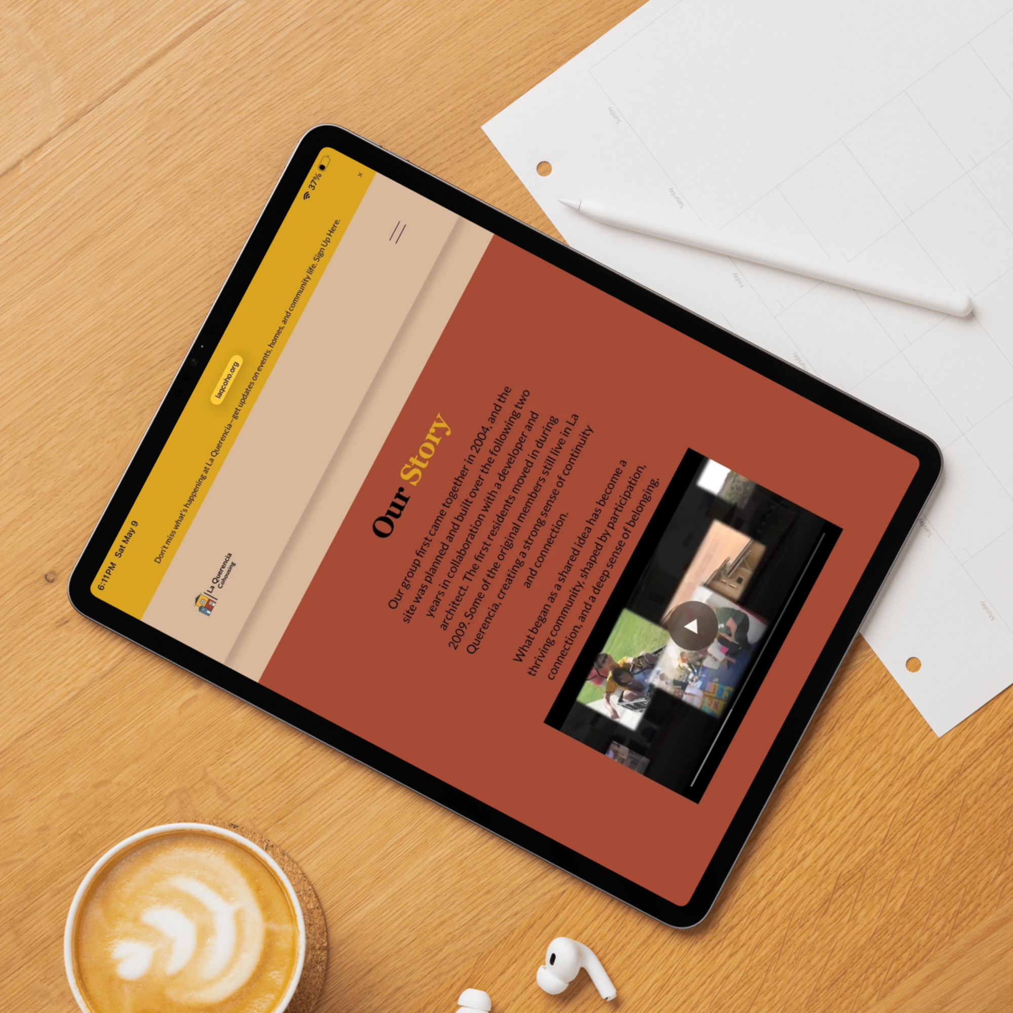 A wooden desk with a coffee cup, earbuds, and a tablet displaying a webpage, with a white sheet of paper and a white pen nearby.