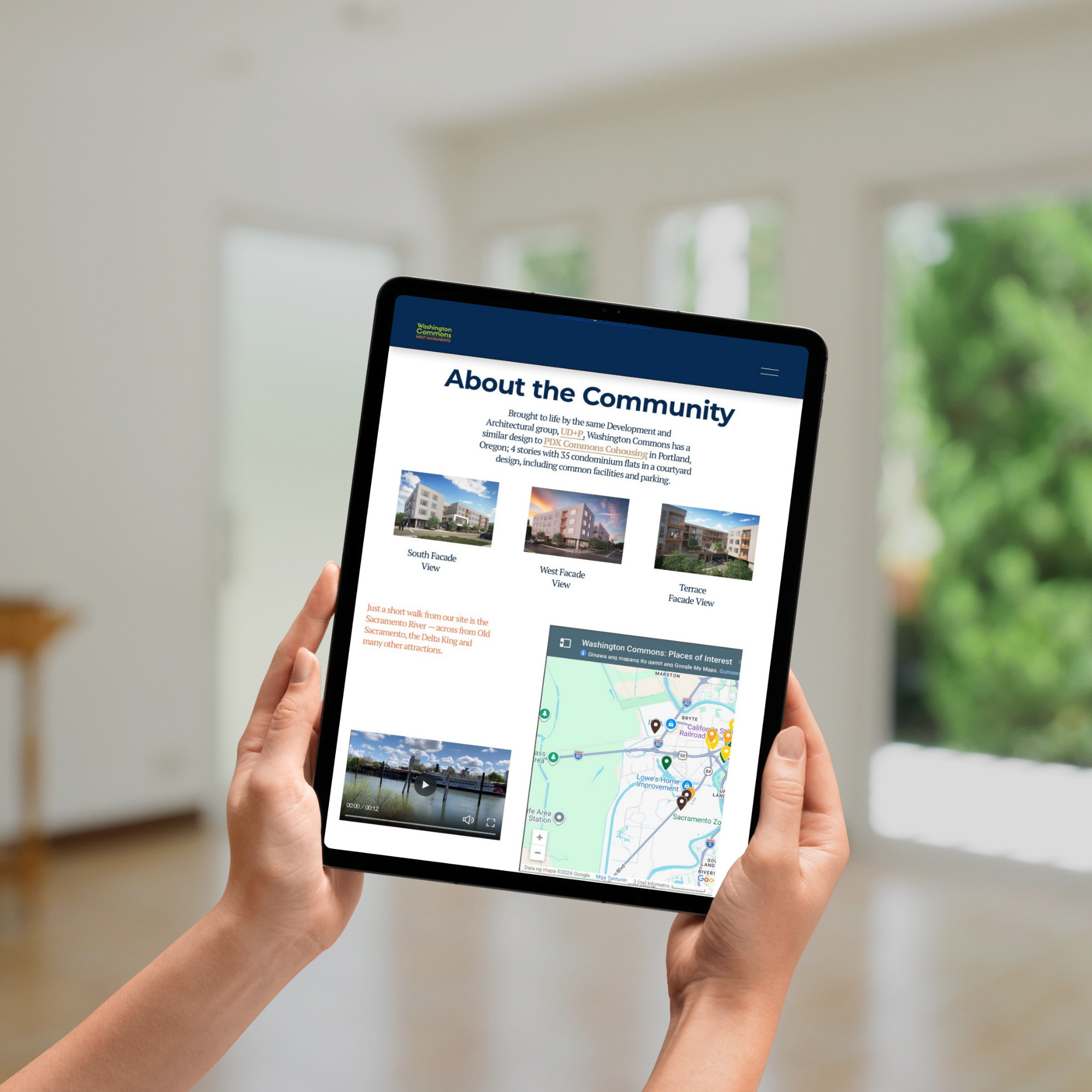 Person holding a tablet displaying a community website with maps and images of buildings, near a window with blurred greenery outside.