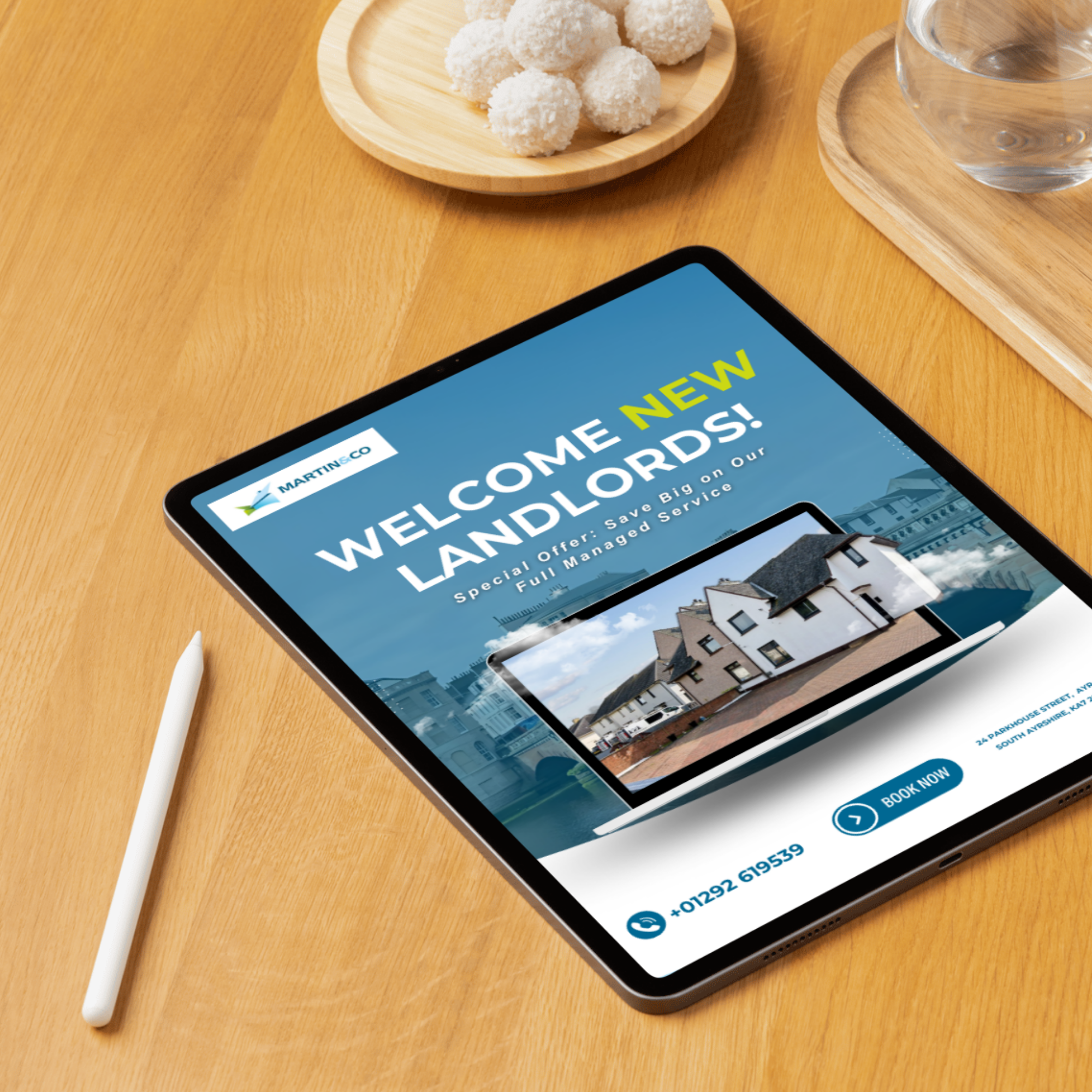 Close-up of a smartphone on a wooden surface displaying a real estate advertisement, with a wooden tray holding white coconut truffles and a glass of water nearby, and a stylus pen beside the phone.