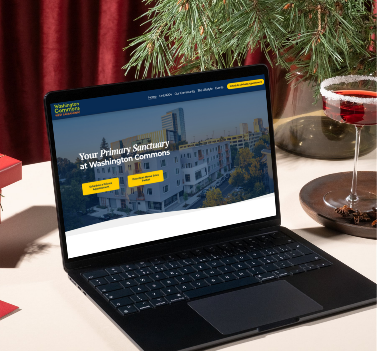 Laptop on a table displaying a webpage for Washington Commons West Sacramento with options to schedule a private appointment or download a home sales packet, next to a glass of red wine and a potted plant.