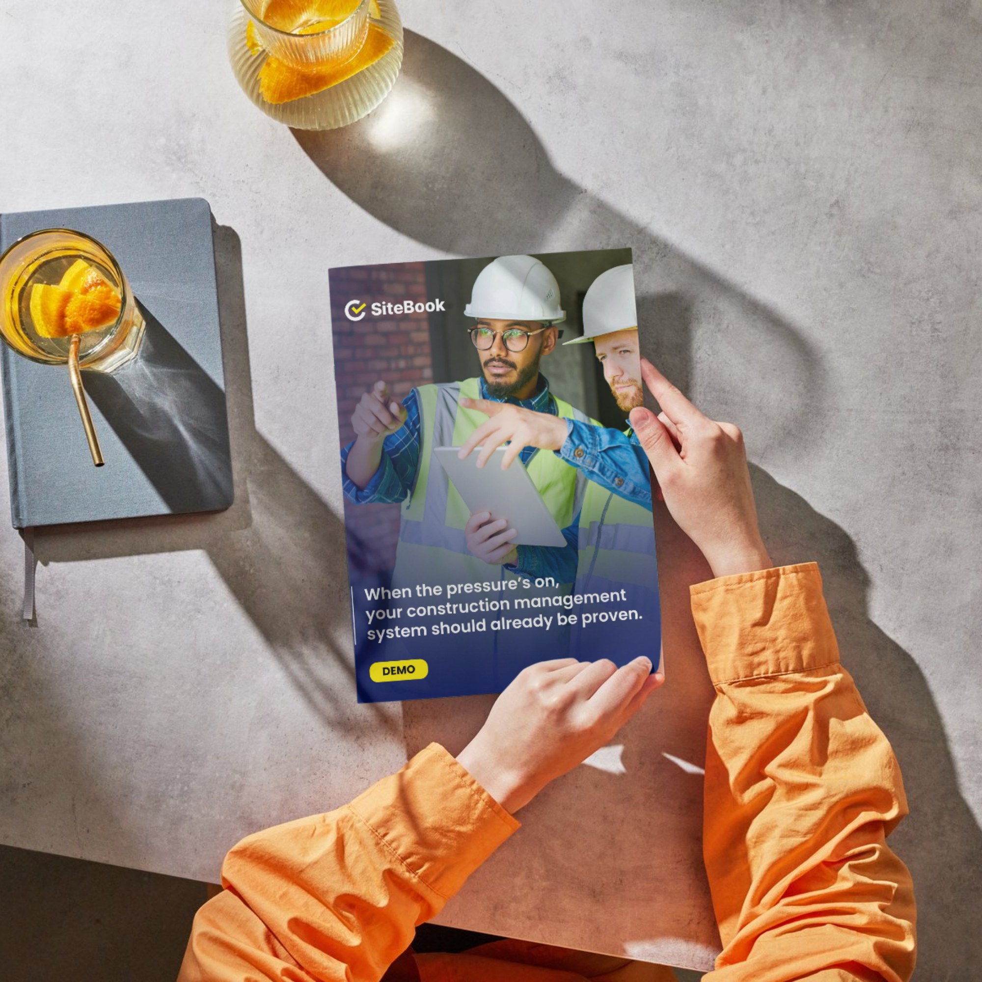A person in orange sleeve holding a brochure with construction workers, a yellow badge with the word 'DEMO,' and text that says 'When the pressure's on, your construction management system should already be proven.' The scene includes two glasses with orange slices and a book on a light-colored table.