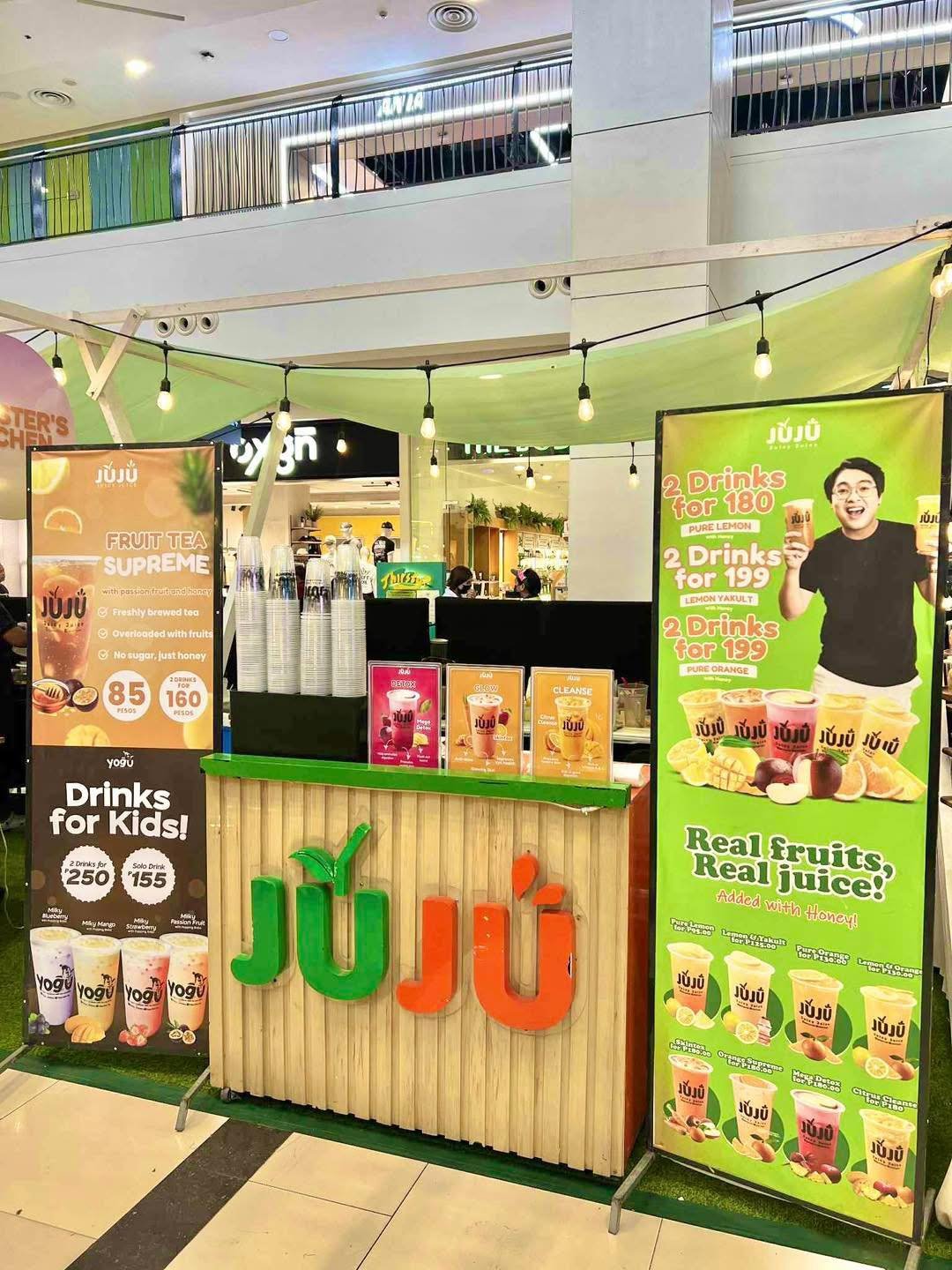 JuJu juice stand in a shopping mall featuring colorful banners with drink deals and images of fruit juices, with a wooden counter displaying paper cups and promotional posters promoting various fruit juice flavors made with real fruits and honey.