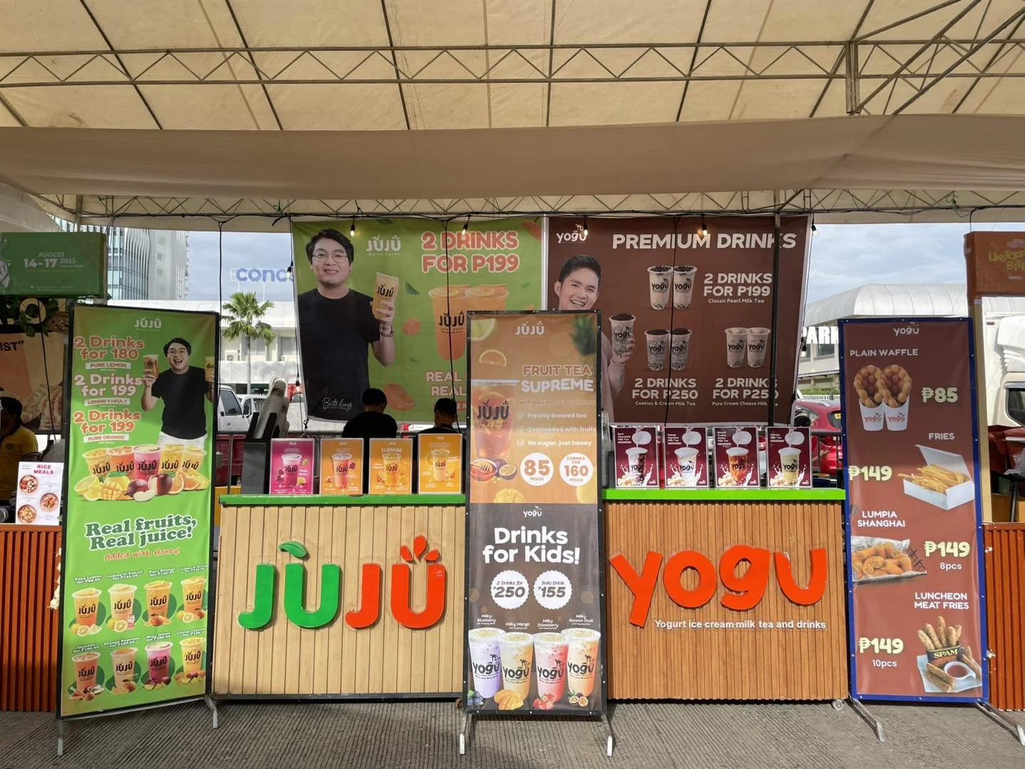 A food stall featuring promotional banners and menu boards for various drinks and snacks, including fruit juices, flavored teas, fries, and fried items, with visible prices and colorful signage.