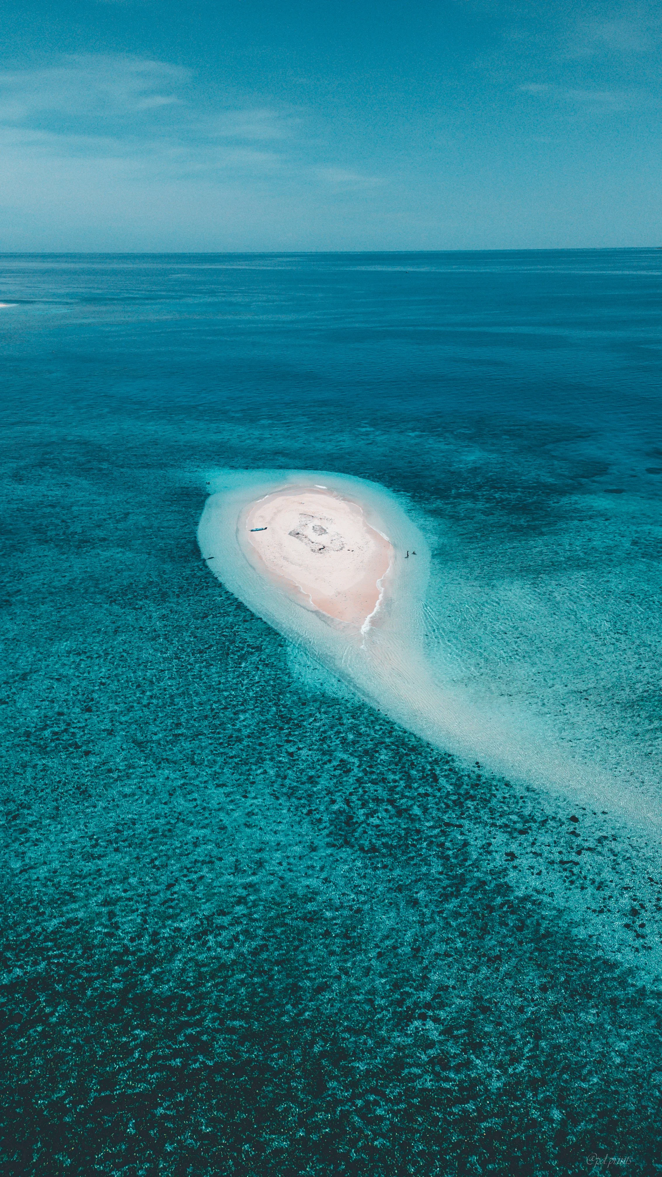 Aerial view of a small, white sandy island with a central structure, surrounded by clear turquoise water and a distant horizon of the ocean under a partly cloudy sky.
