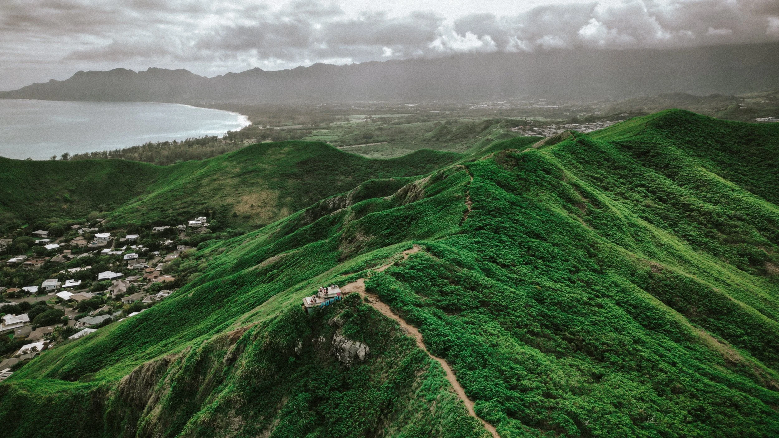 Hiking trail on lush green hills with a town below and mountains in the distance under cloudy skies.