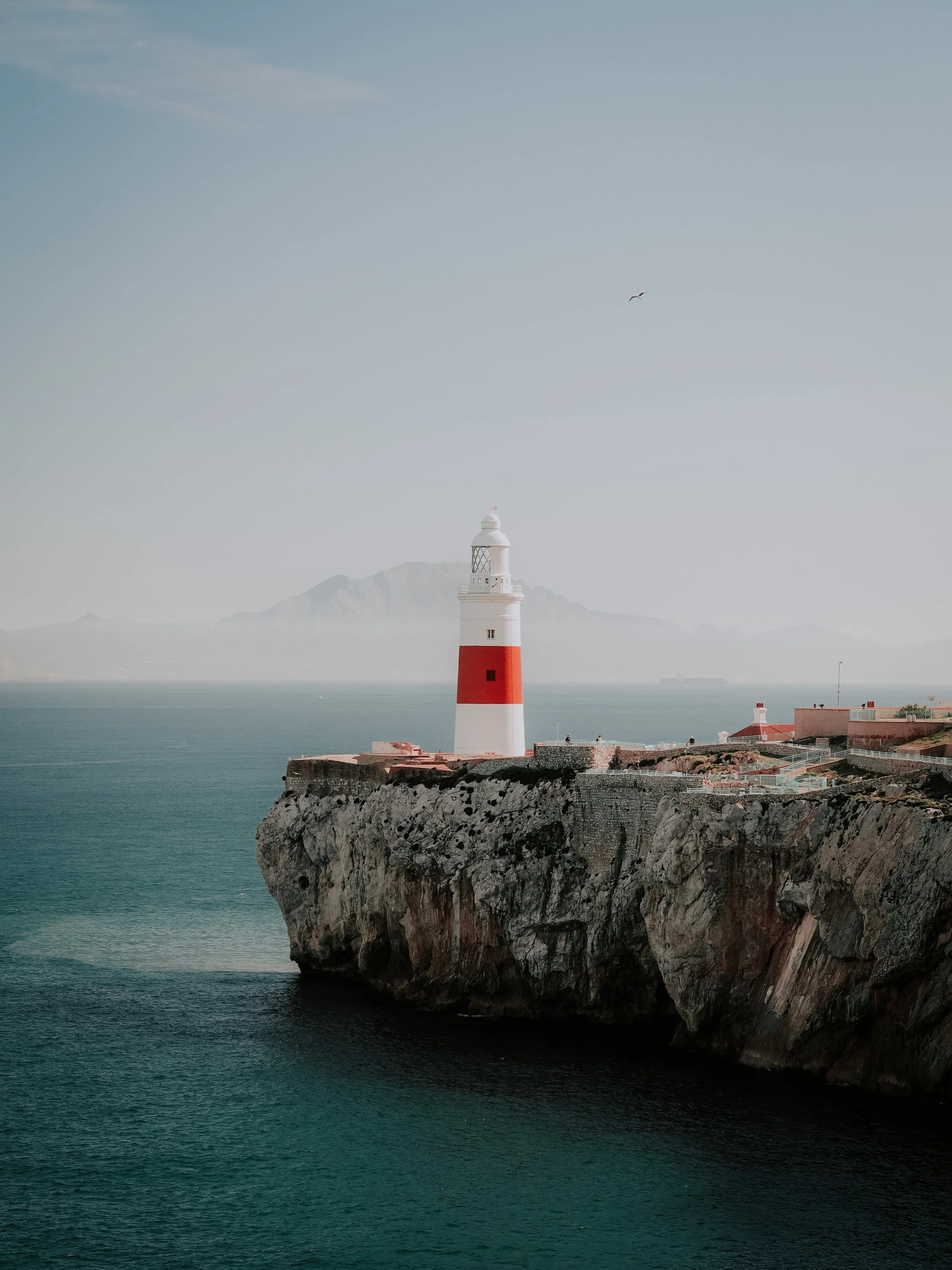 A lighthouse on a rocky cliff with a mountain in the background and a bird flying in the sky.