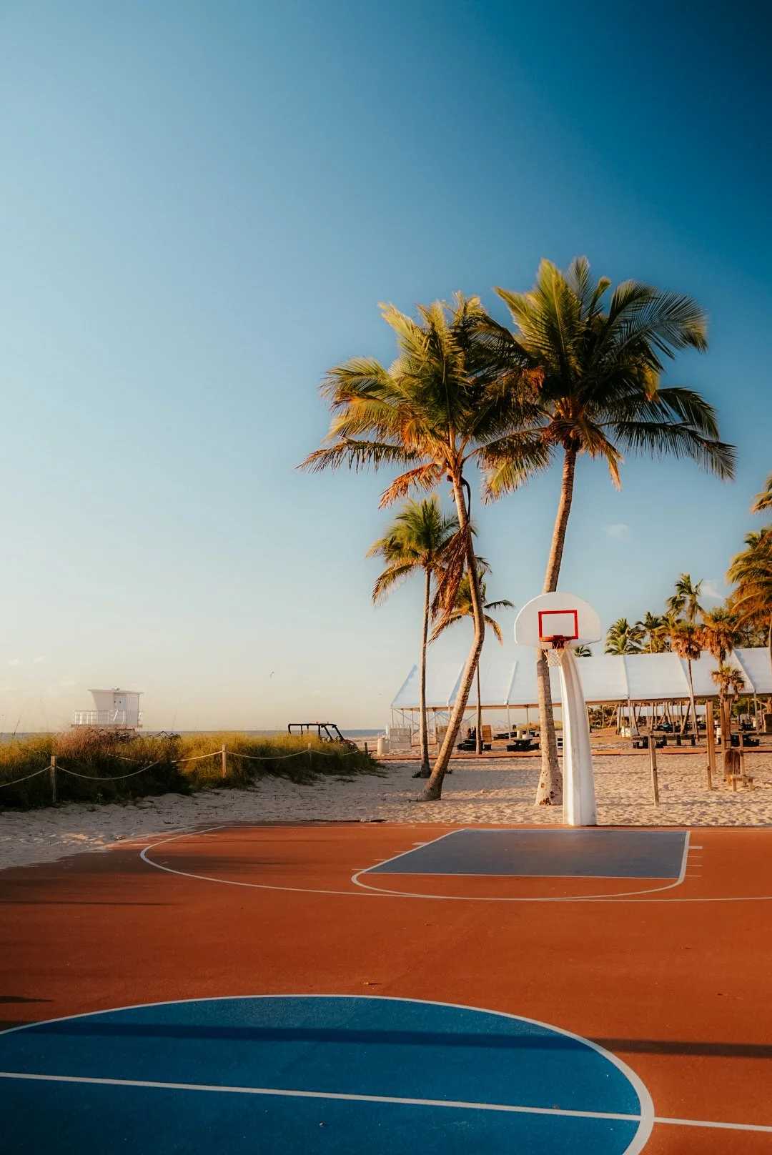 A beachside basketball court with palm trees, sand, and beach tents in the background during sunset.