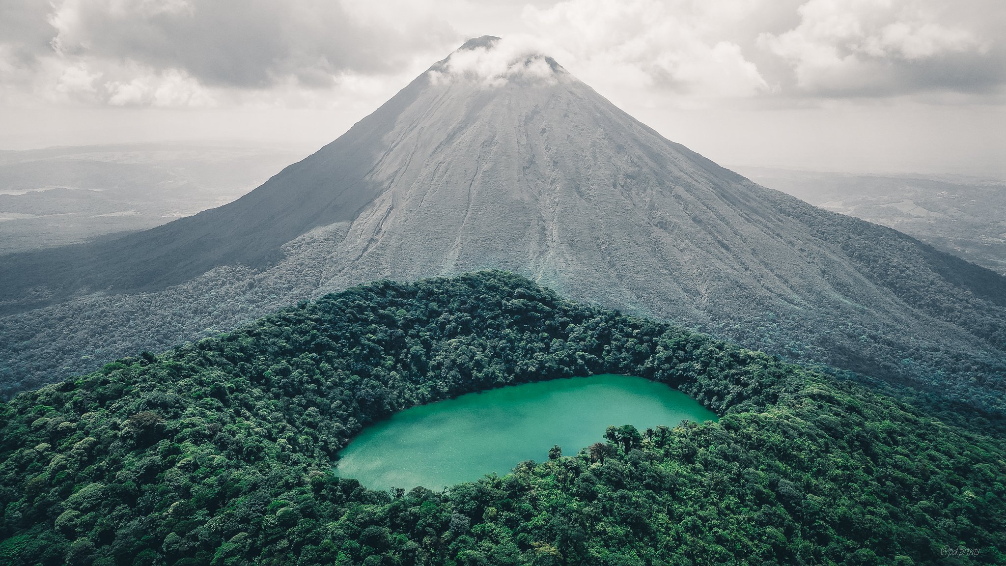 A large, conical volcano with a gray, rocky surface and patches of clouds near the top. In the foreground, there is a lush green forest surrounding a small, turquoise crater lake.