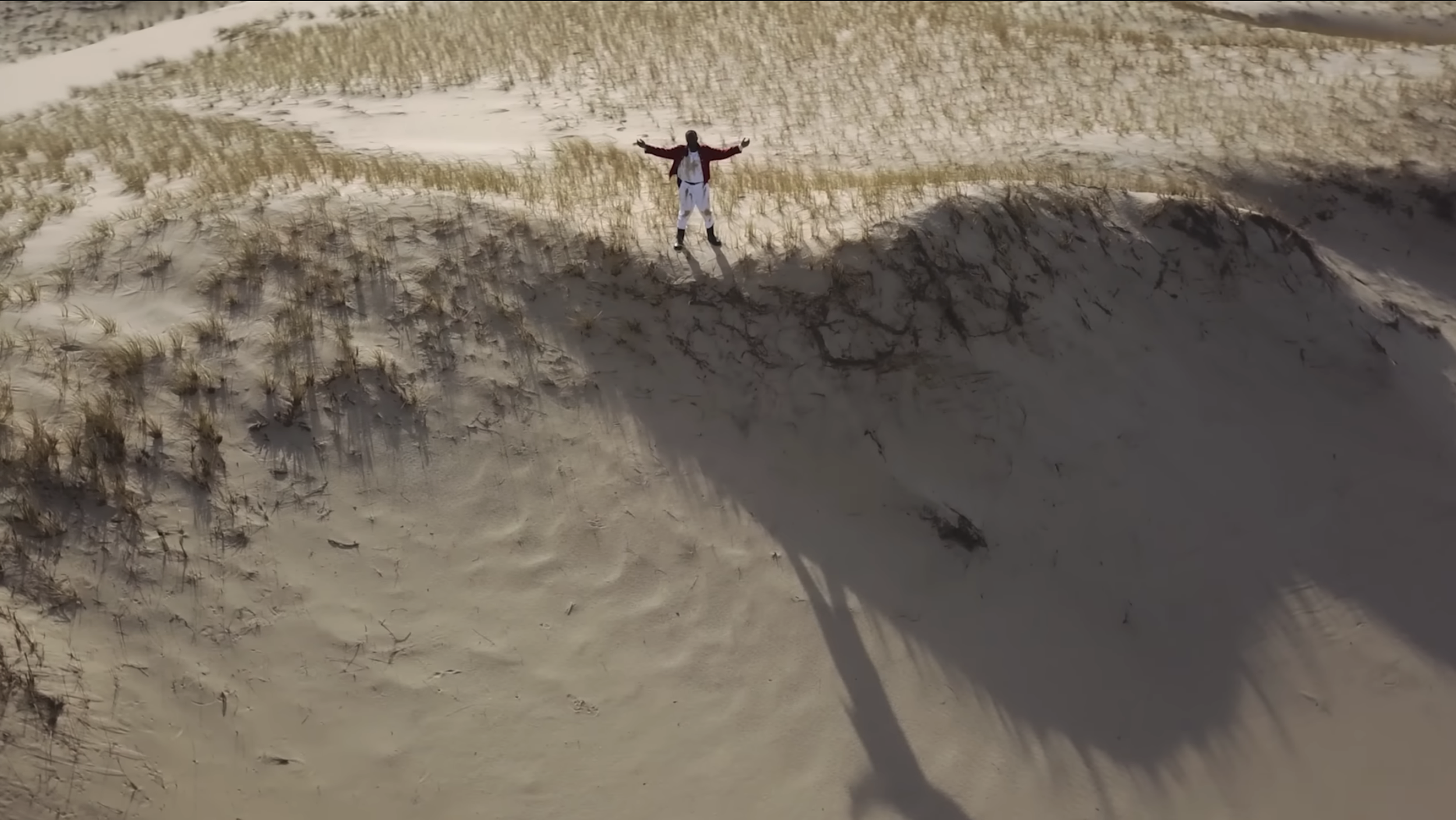 A person standing on sandy dunes with arms outstretched, surrounded by patches of grass and casting a shadow.