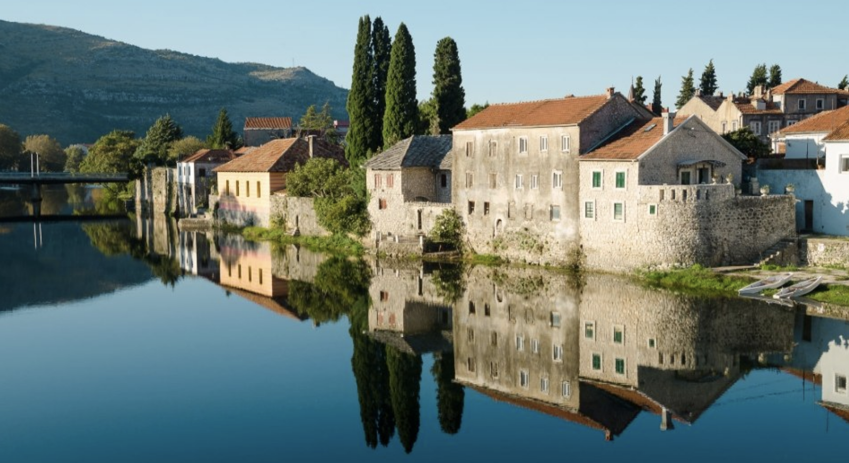 A peaceful riverside village with old stone houses, tall cypress trees, and mountains in the background. The water reflects the houses and trees.