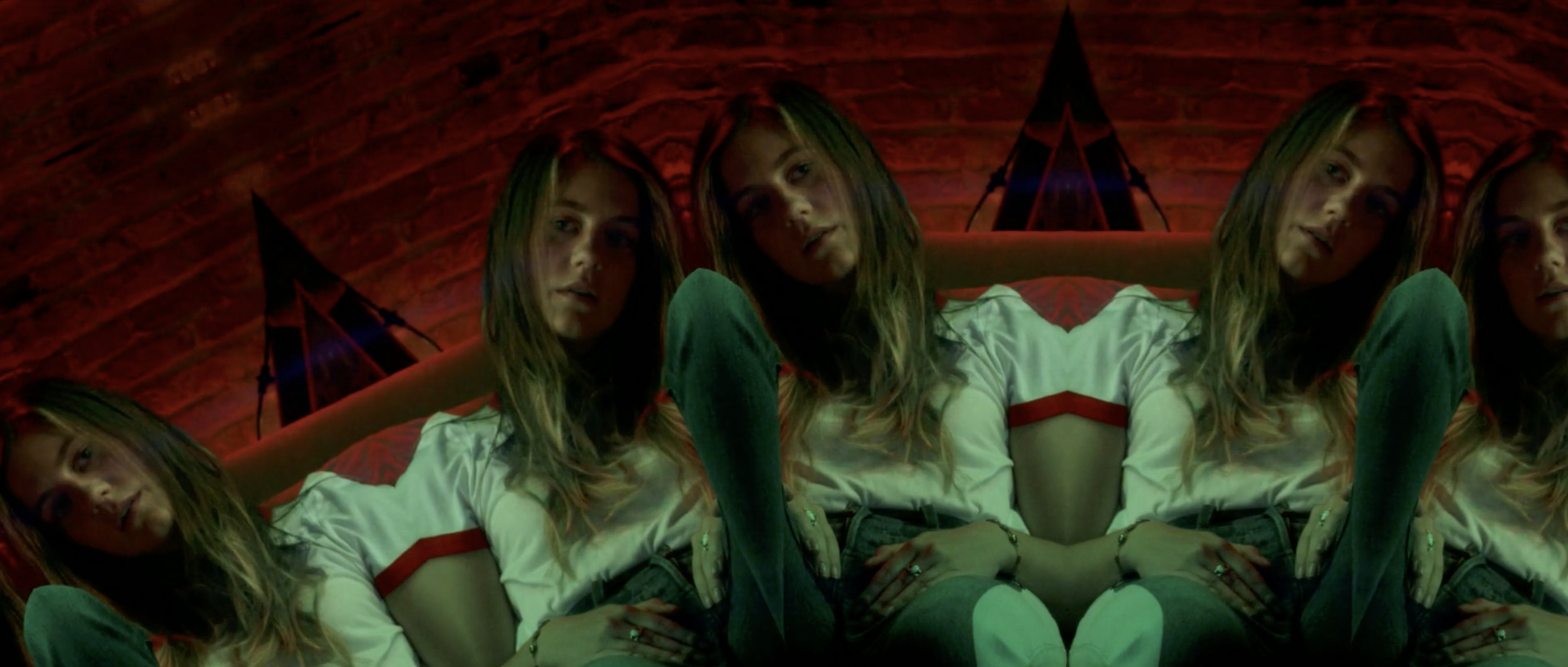 A distorted, mirrored image of a woman with long blonde hair sitting on a bed with a green pillow, wearing a white t-shirt with red stripes, and a black bracelet, in a dimly lit room with a red brick wall and black star-shaped wall decorations behind her.