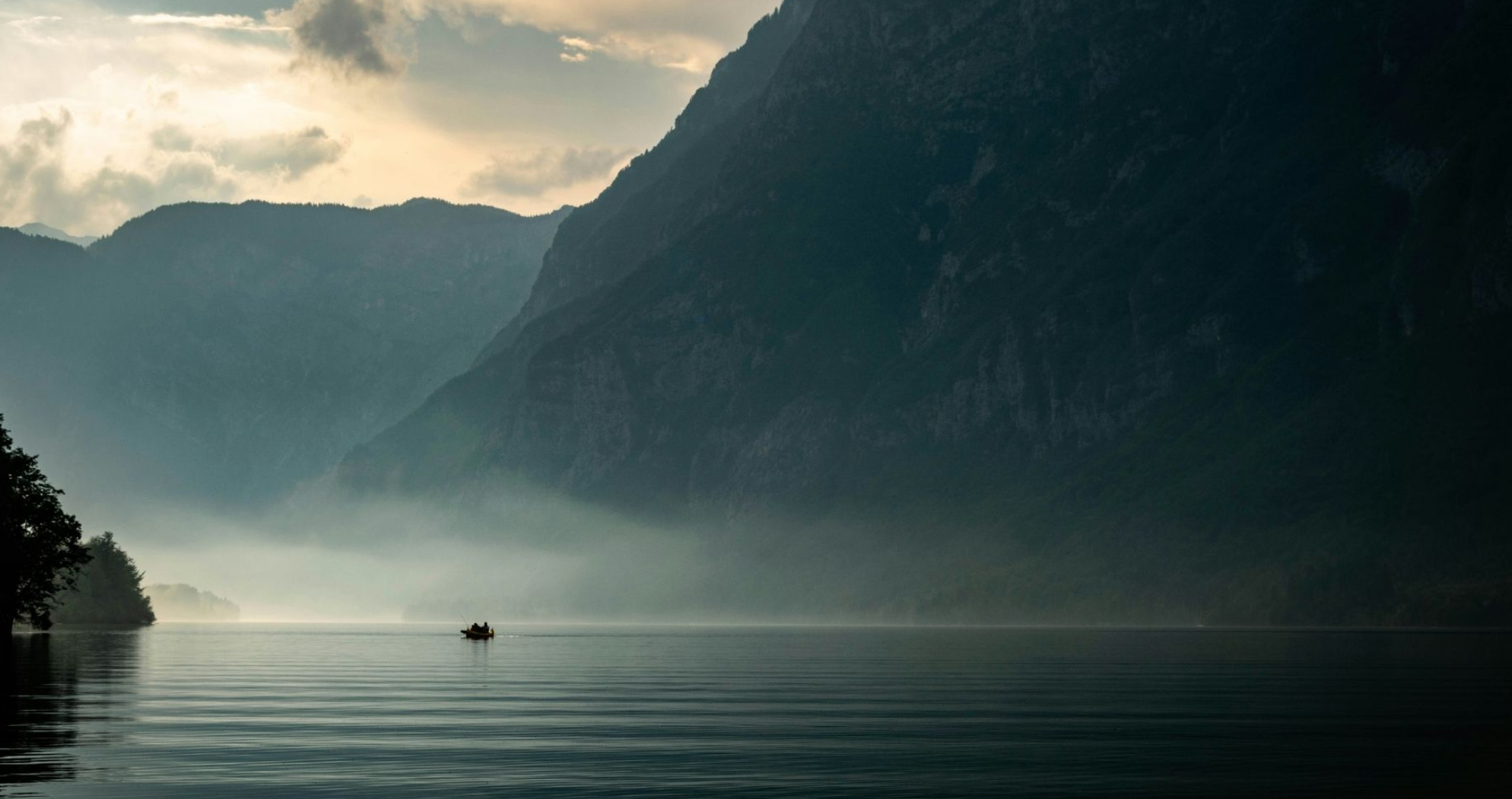 A boat floating on a calm lake surrounded by tall mountains and mist, with cloudy sky overhead.