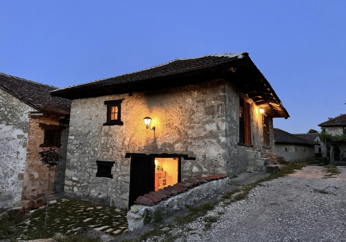 A rustic stone house with a wooden roof and small windows, illuminated by warm indoor and outdoor lights, during twilight time.