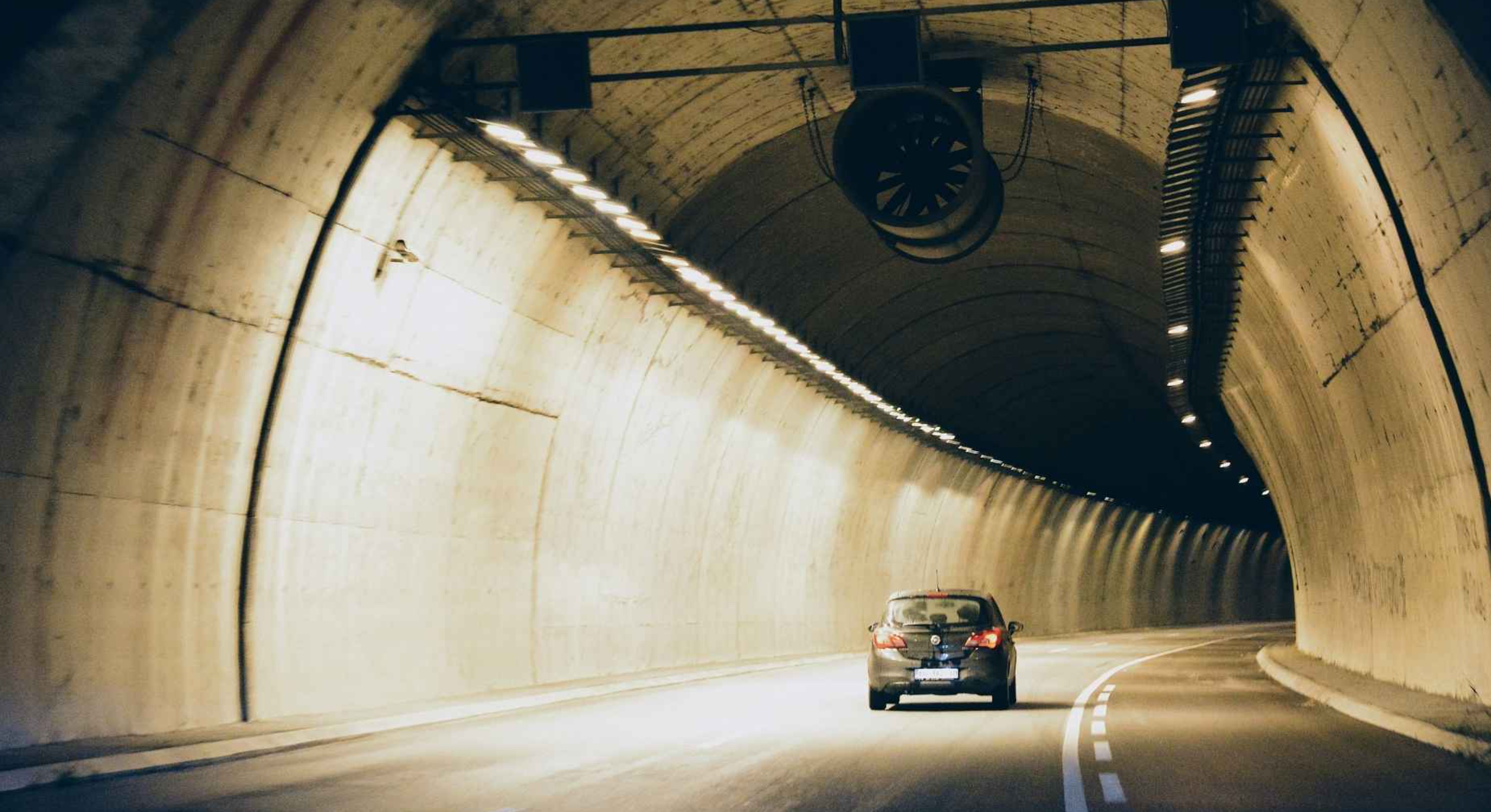 A car driving through a tunnel with curved concrete walls and ceiling, illuminated by line of lights along the top edge and machinery hanging from the ceiling.